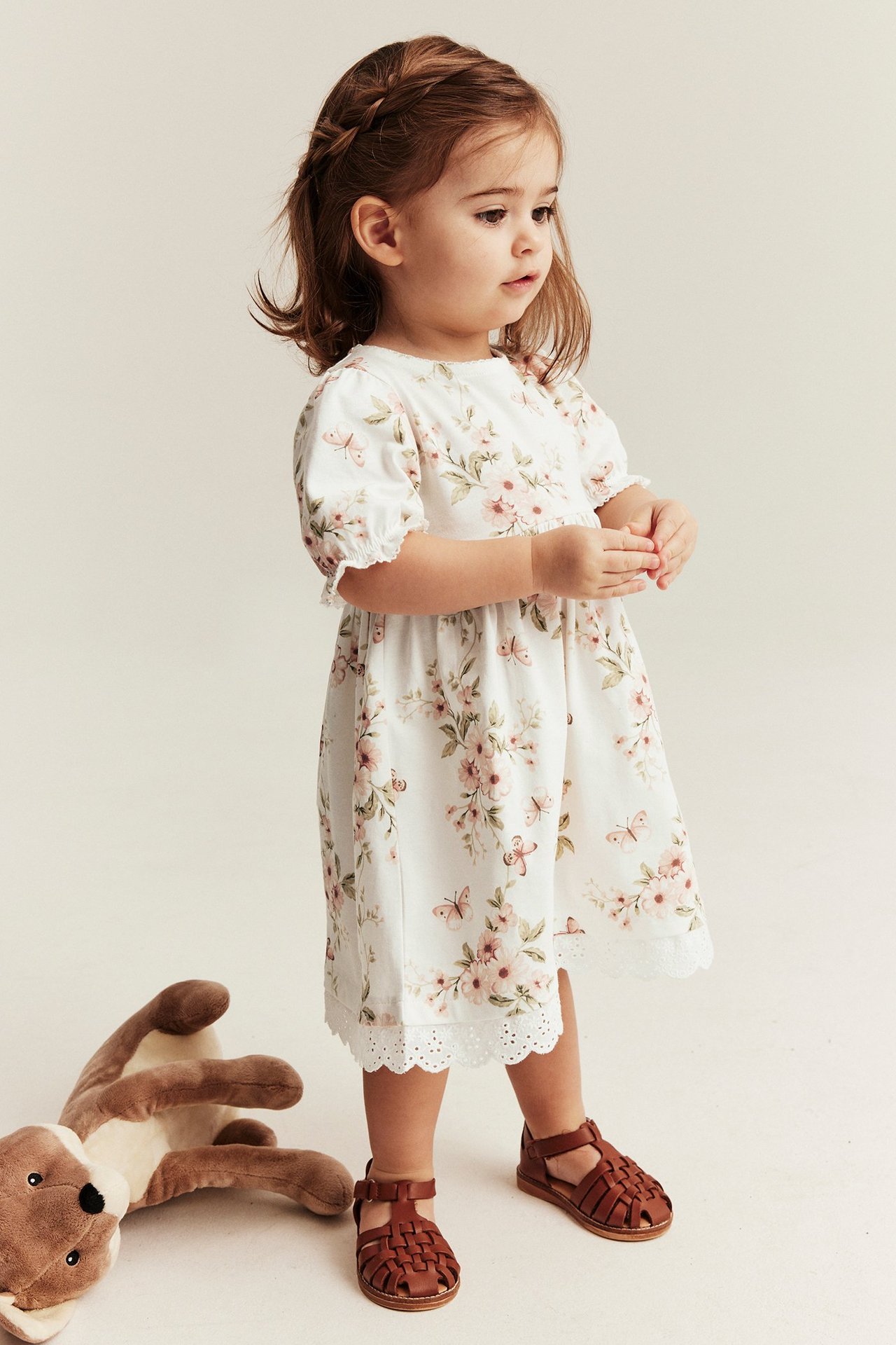 Widok z przodu: Dziecko w kremowej bawełnianej sukience w kwiaty i motyle z bufiastymi rękawami oraz brązowych sandałach.
