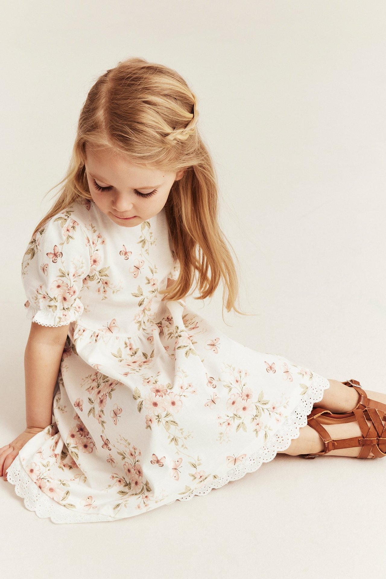 Dziewczynka w ecru kwiecistej bawełnianej sukience z bufiastymi rękawami, koronką i brązowymi sandałami, widok z przodu.