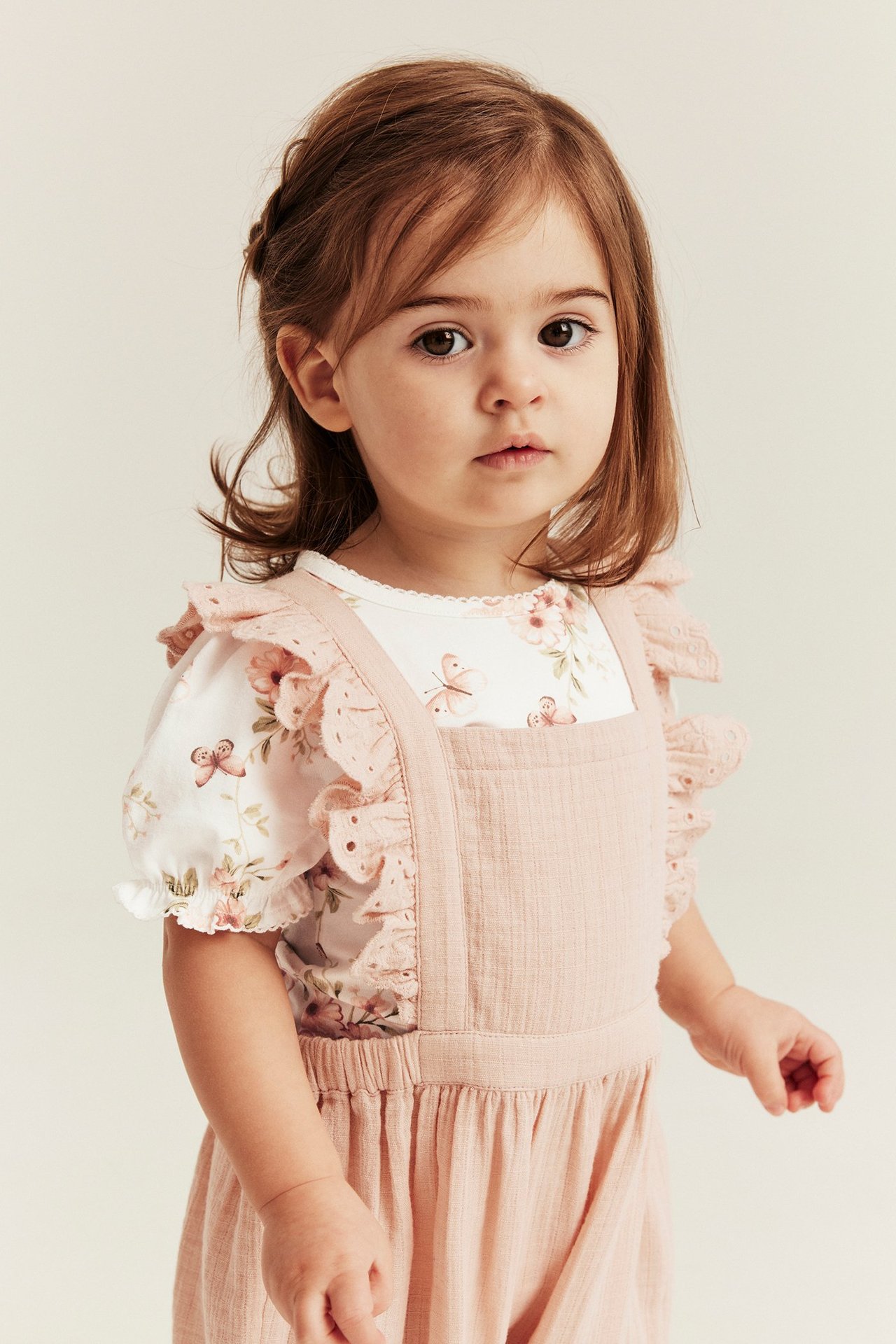 Z przodu: dziecko w kremowym body w kwiaty z bufiastymi rękawami pod różowym kombinezonem z falbanami.