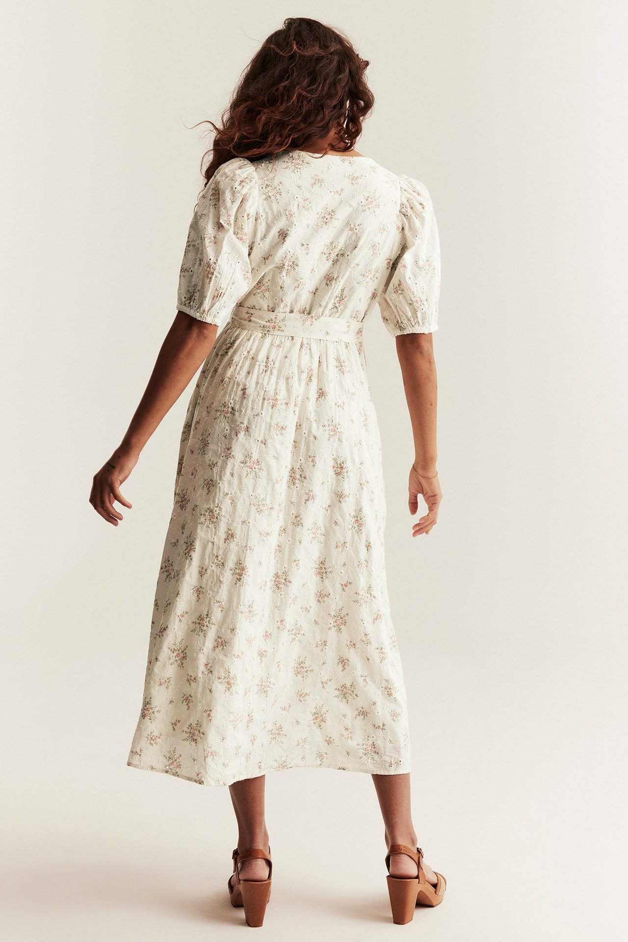Bakfra: kvinne i offwhite bomullskjole med broderte blomster, puffermer og brune sandaler.