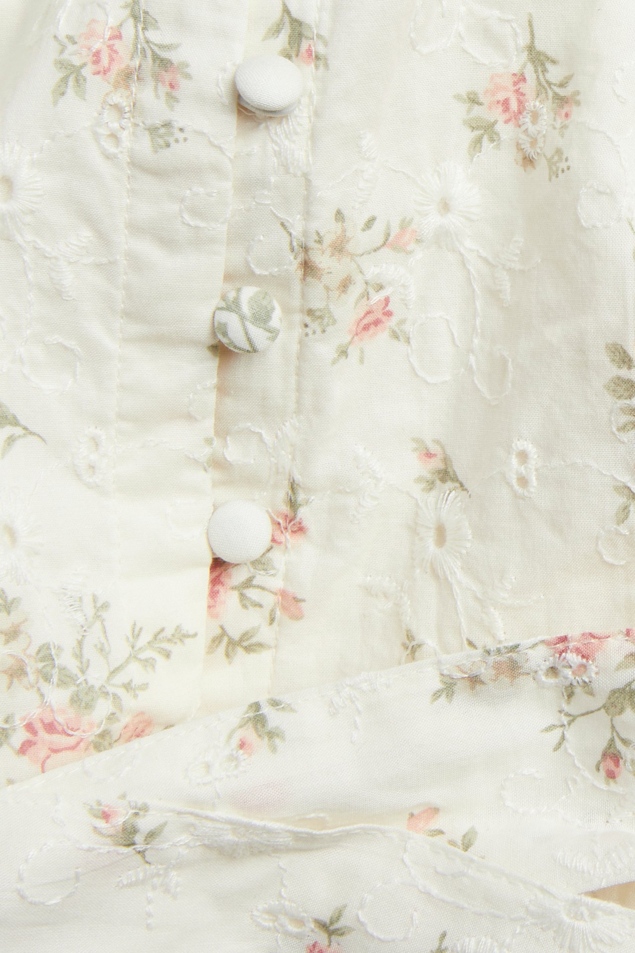 Nærbilde av en offwhite bomullskjole med rosa blomstermønster, broderte detaljer og stofftrukne knapper foran.