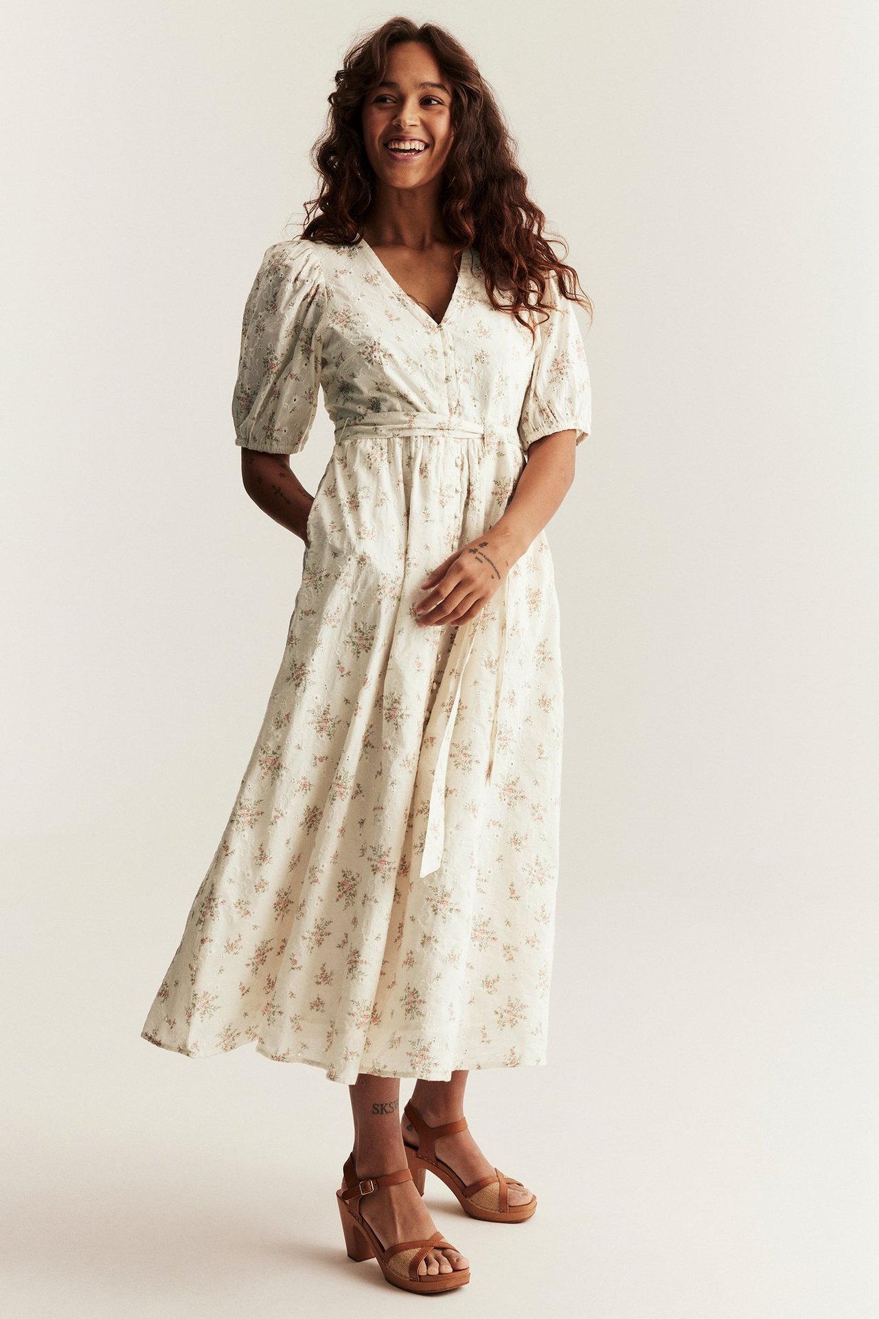 Frontbilde av en kvinne i en offwhite blomstret bomulls midikjole med puffermer og brune blokkhæl-sandaler.