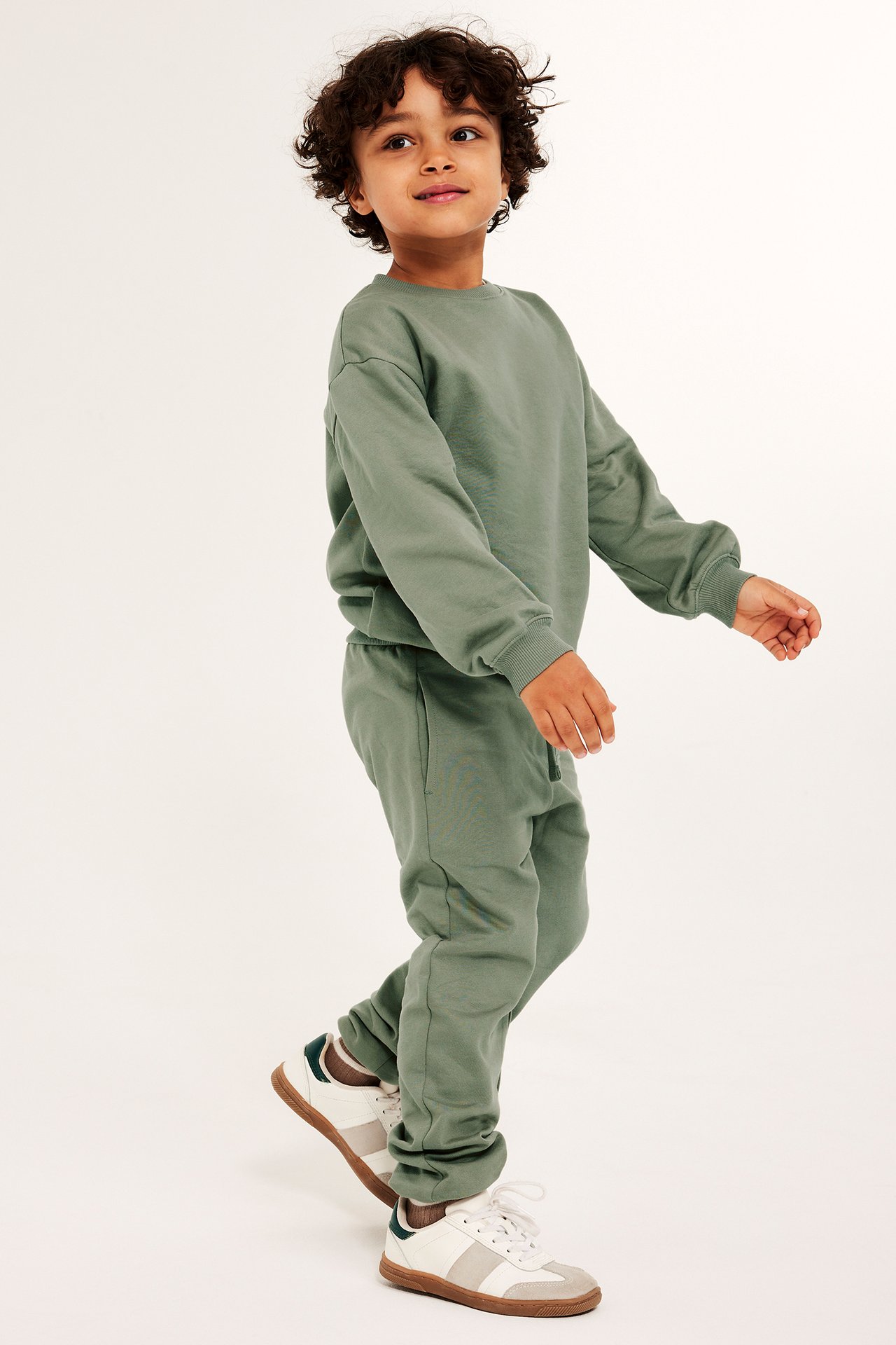 Widok z przodu dziecka w ciemnozielonej bawełnianej bluzie dresowej i pasujących spodniach z białymi trampkami.