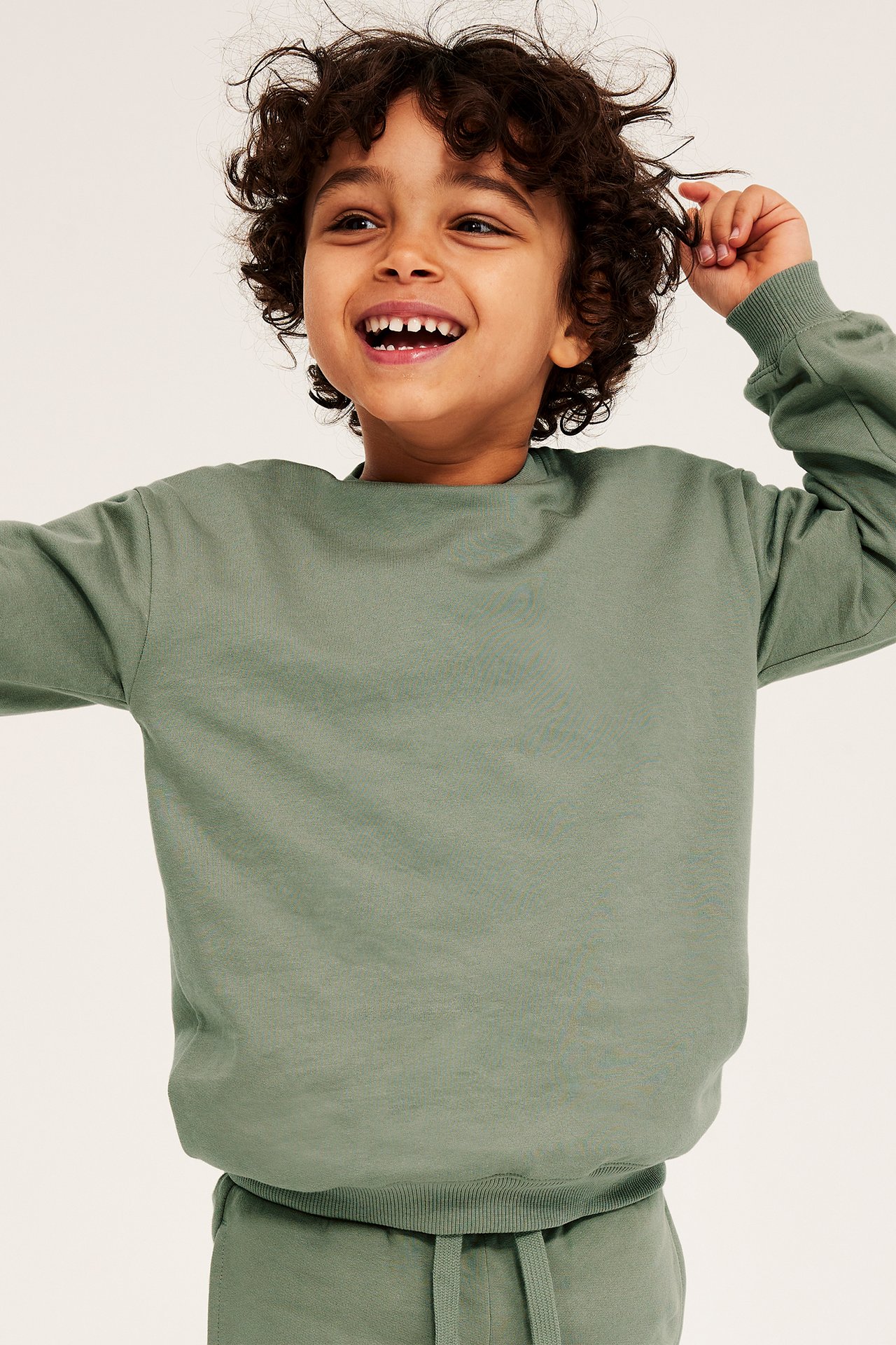 Widok z przodu uśmiechniętego chłopca w ciemnozielonej bawełnianej bluzie z prążkowanymi detalami i pasujących spodniach dresowych.