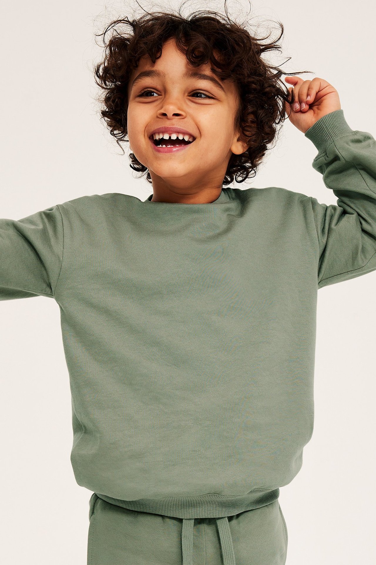 Widok z przodu uśmiechniętego chłopca w ciemnozielonej bawełnianej bluzie z prążkowanymi detalami i pasujących spodniach dresowych.
