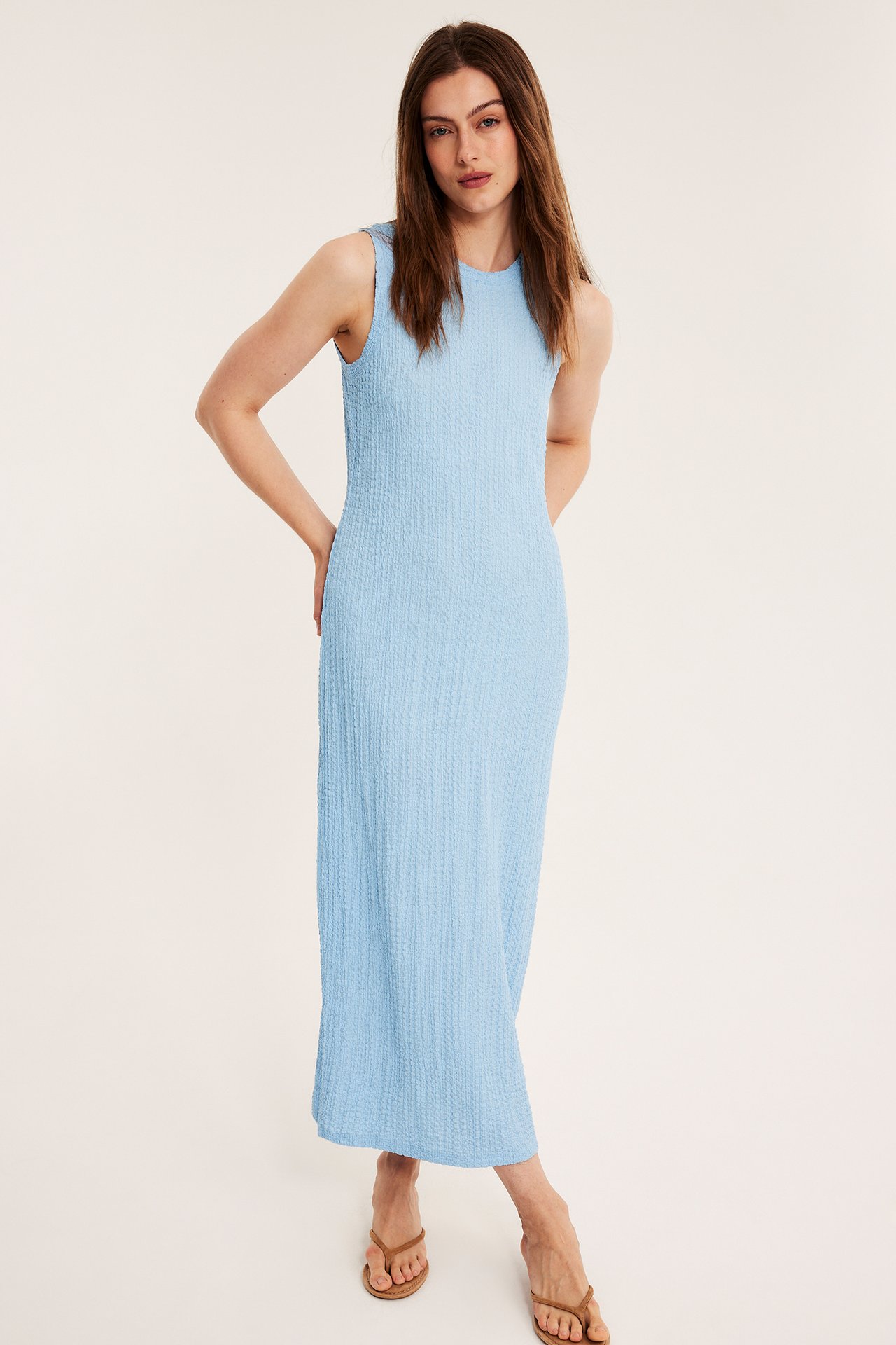 Framifrån, kvinna i ljusblå ärmlös midiklänning med strukturerat tyg och bruna platta sandaler.