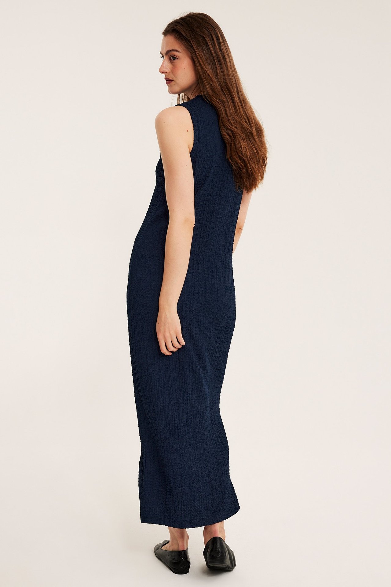 Naisen takasivukuva tummansinisessä hihattomassa tekstuurisessa midimekossa ja mustissa ballerinoissa.