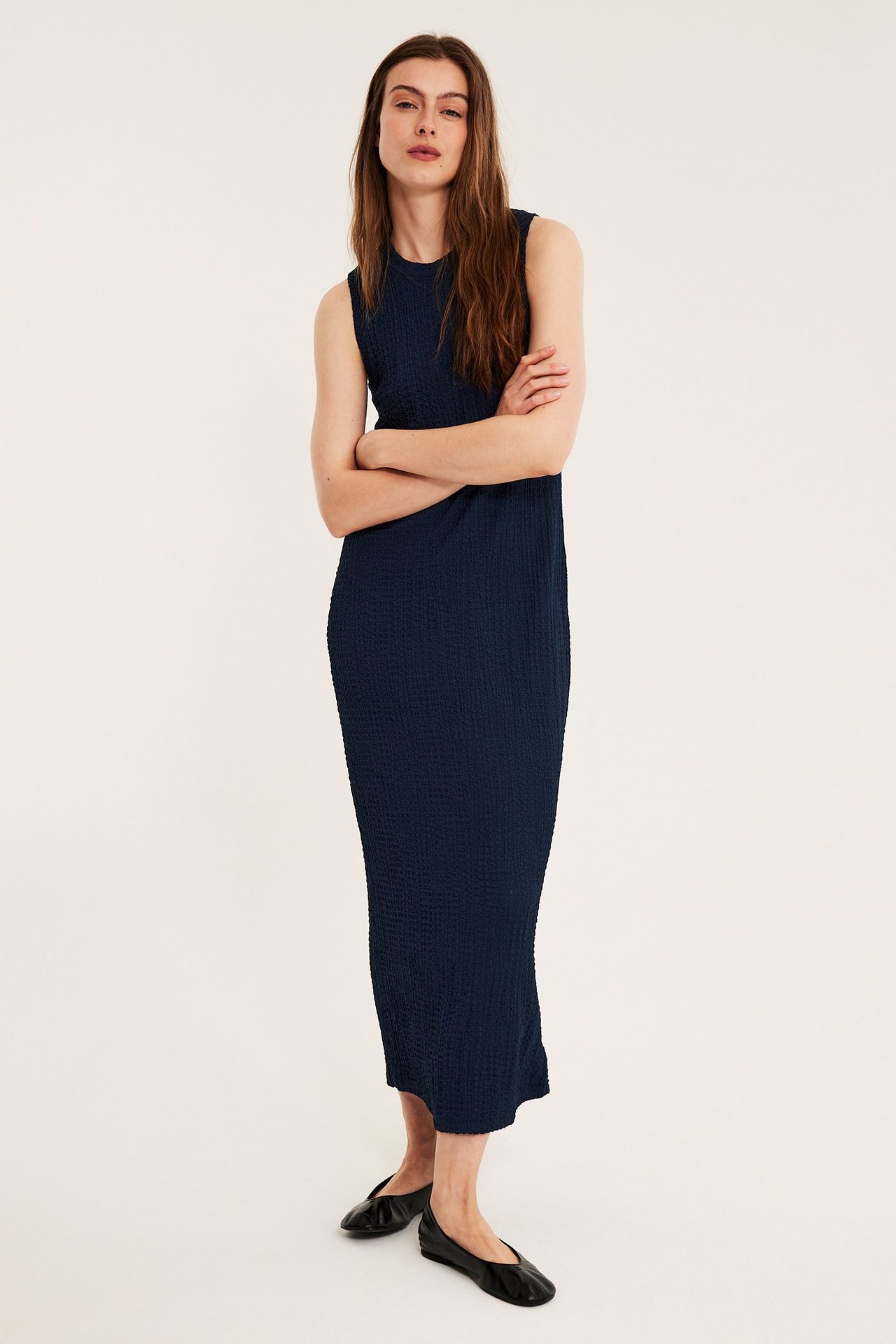 Edestäpäin, nainen yllään tummansininen hihaton strukturoitu midimekko ja mustat ballerinakengät.