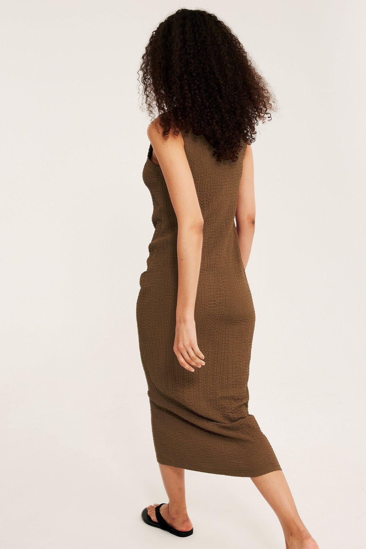 Naisen takakuva yllään istuva ruskea strukturoitu hihaton midimekko ja mustat litteät sandaalit.