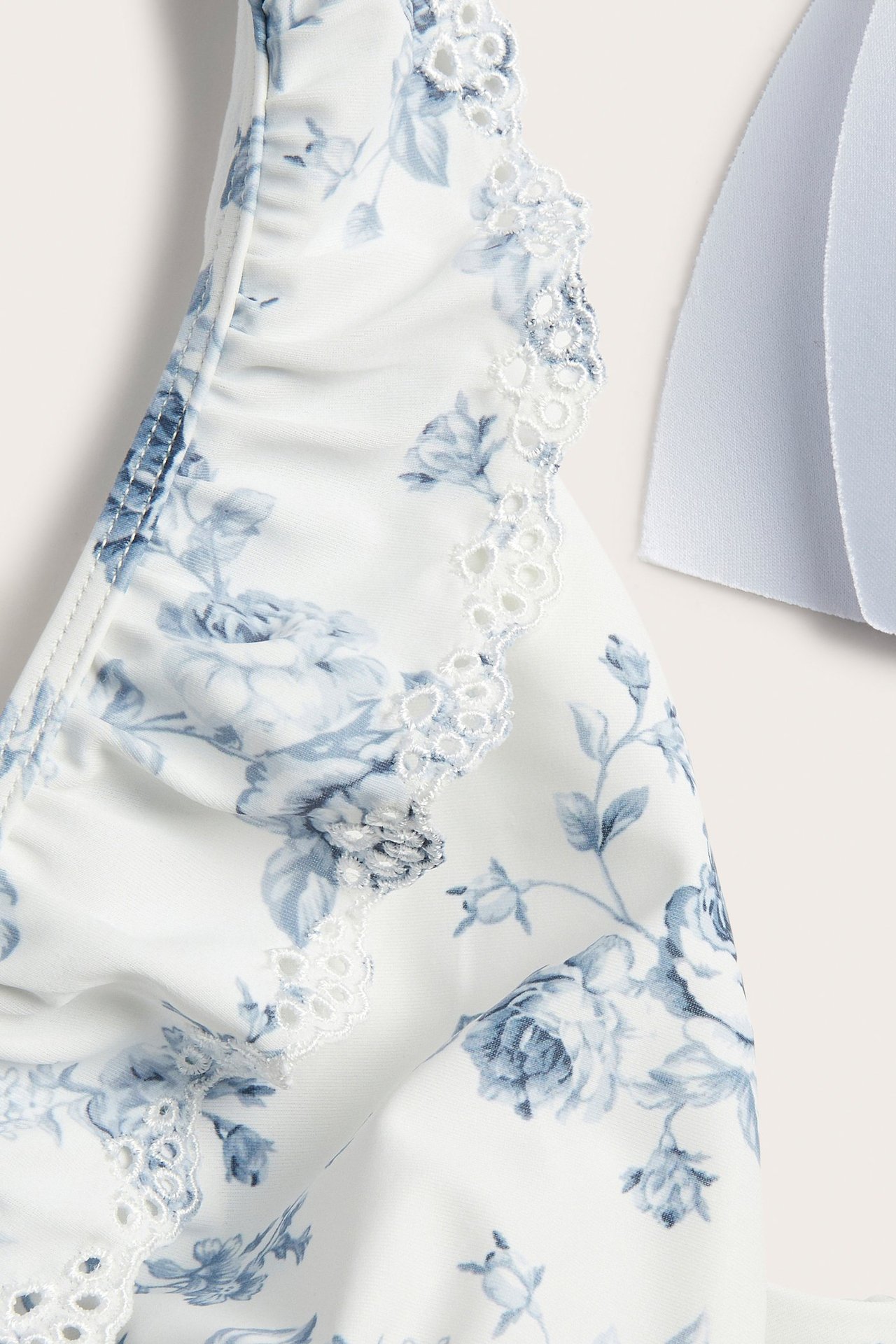 Zbliżenie na kremowy kwiecisty kostium kąpielowy z niebieskim wzorem i białą, haftowaną falbanką na ramieniu.