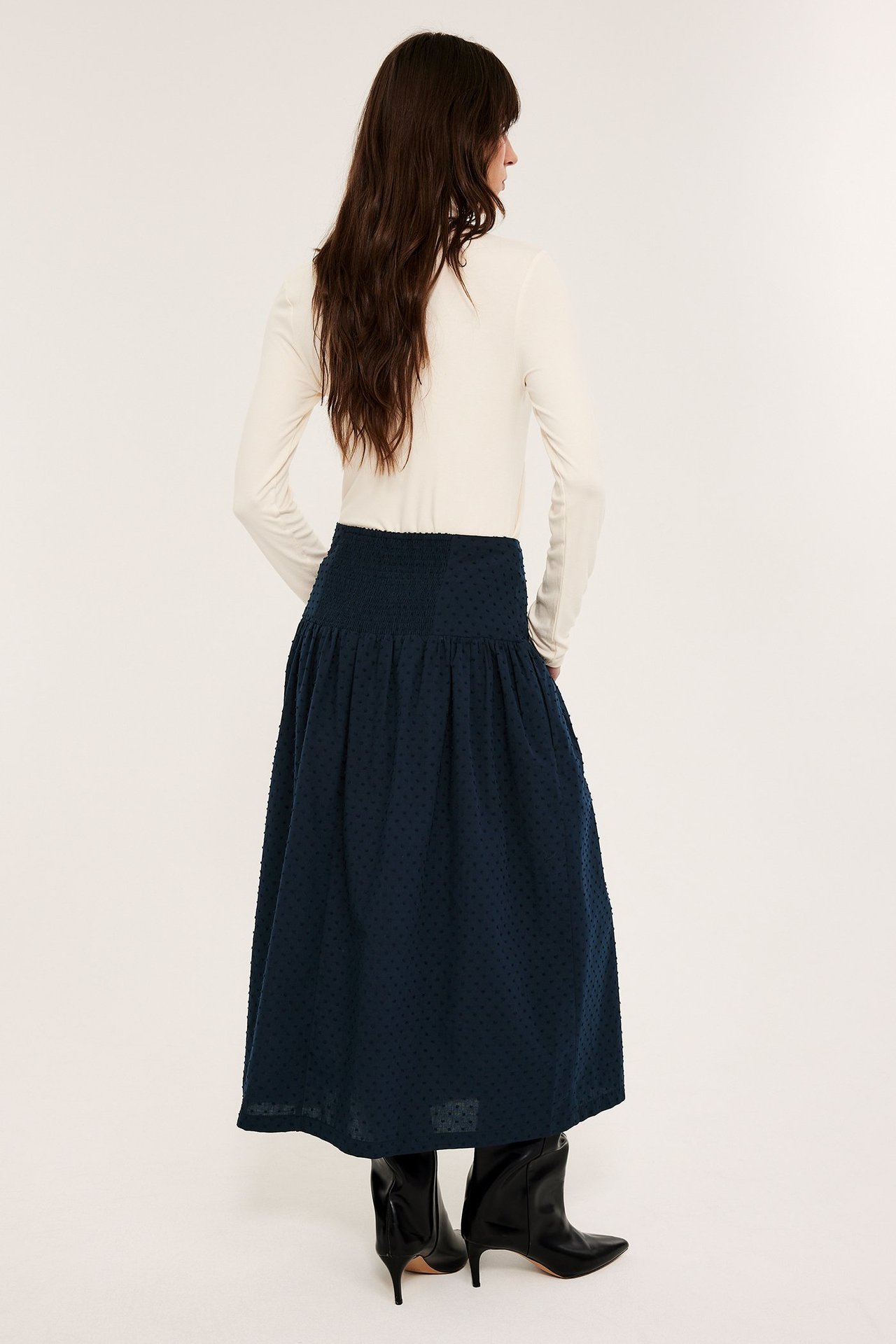 Bakfra: kvinne i mørkeblått prikkete bomulls-midiskjørt, kremfarget ribbet topp og svarte støvler.