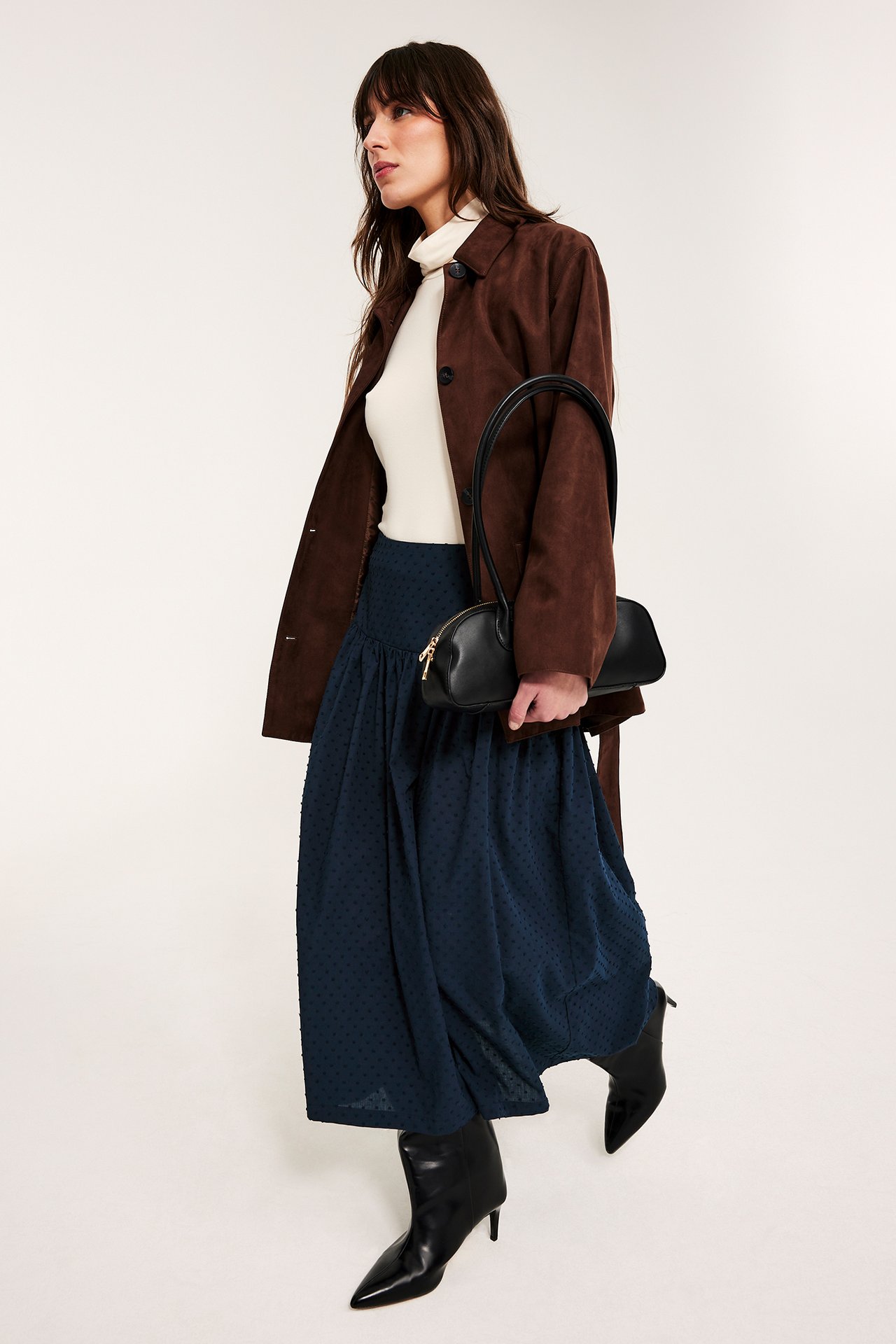 Frontvisning: Kvinne i mørkeblått prikkete midiskjørt, beige genser, brun jakke, svarte støvler, svart veske.