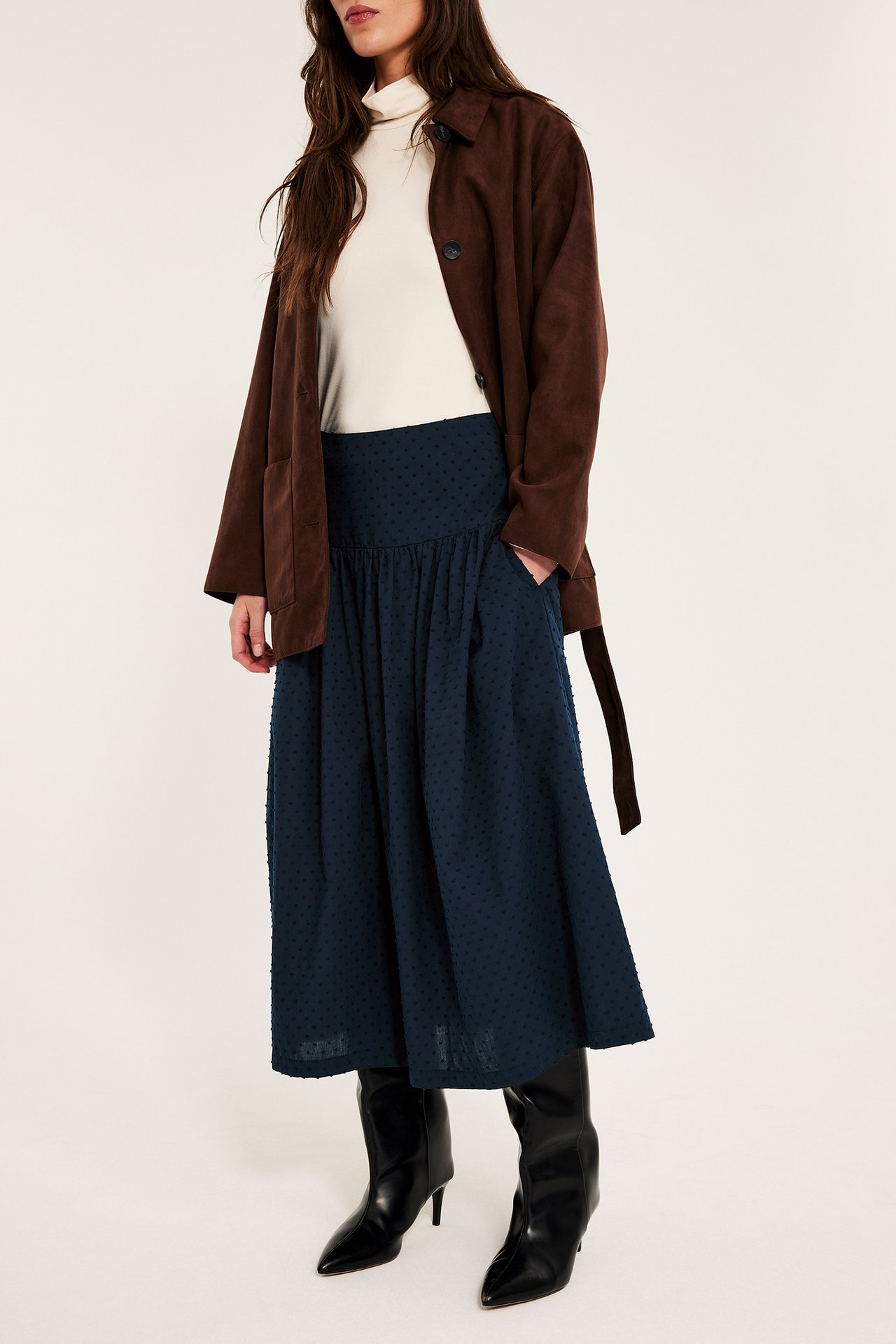 Frontutsikt: kvinne i mørkeblått prikkete bomulls-midiskjørt, kremfarget pologenser, brun jakke og svarte hælstøvler.