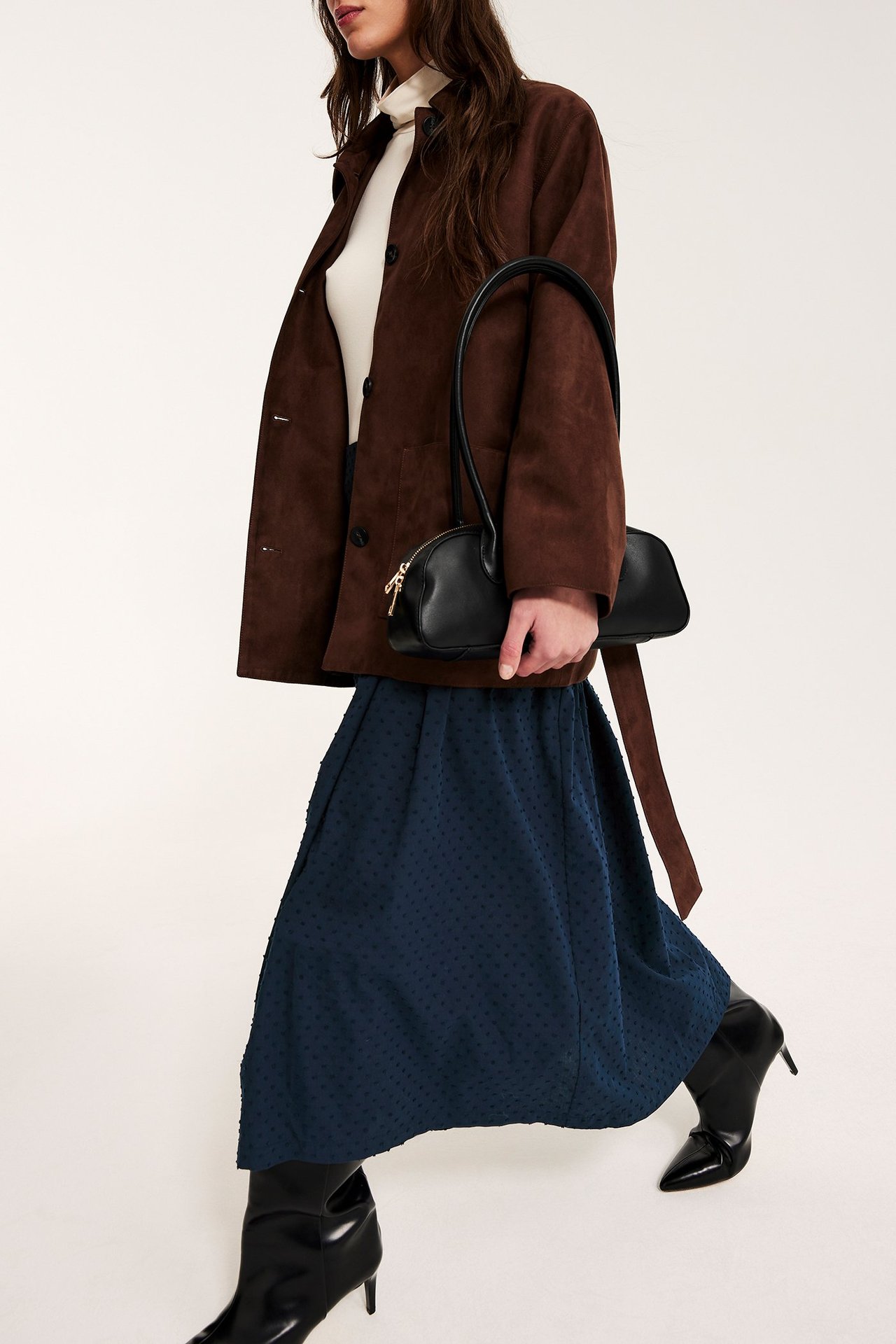 Sidebilde: Kvinne i mørkeblått prikket midiskjørt, brun jakke, hvit topp, svart veske, svarte ankelstøvler.