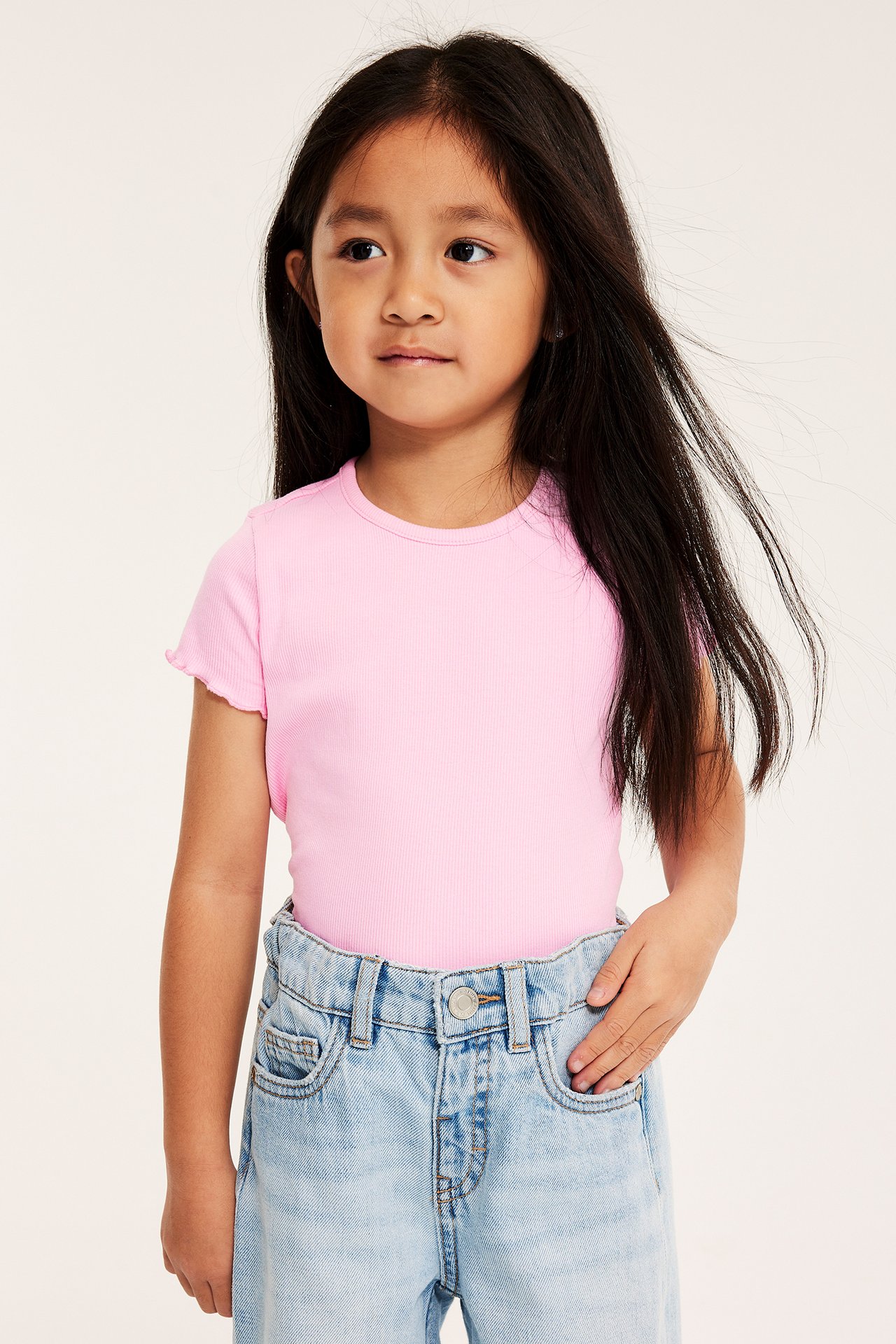 Frontbilde av et barn i rosa ribbet kortermet topp med babylockkanter og lyseblå jeans.