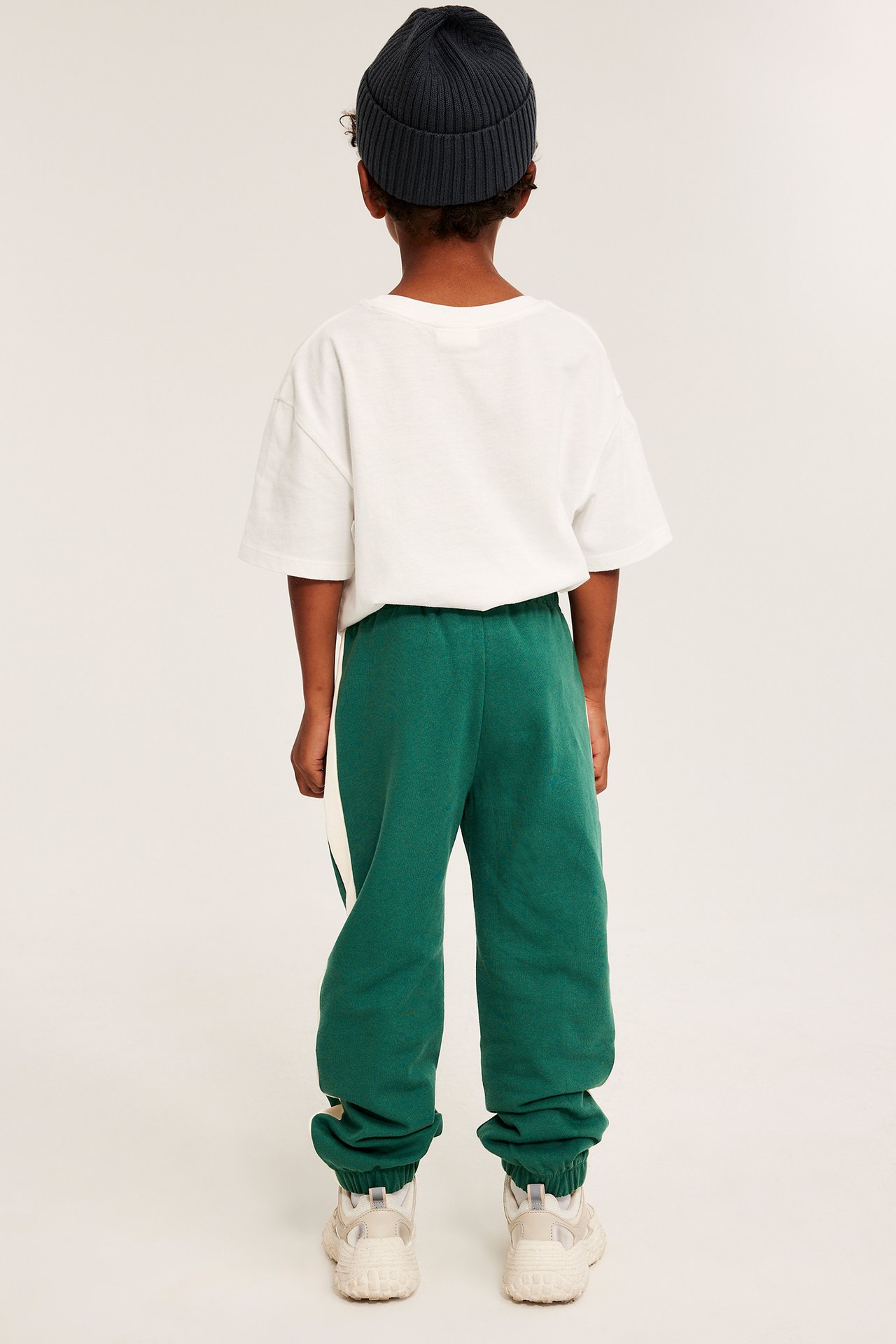 Lapsen takakuva: vihreät puuvillahousut valkoisilla sivuraidoilla, valkoinen t-paita, tummanharmaa pipo, vaaleat lenkkarit.