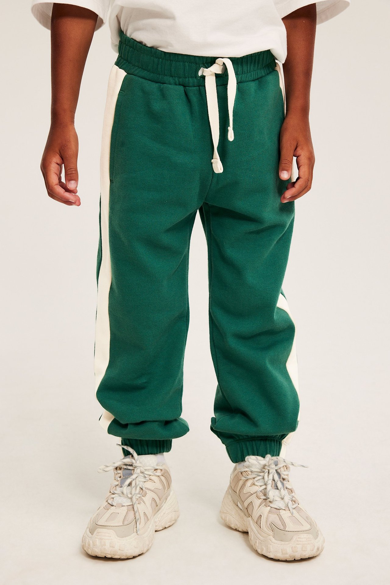 Frontutsikt: barn i grønn joggebukse med kremfargede sidestriper, hvit t-skjorte, beige joggesko.