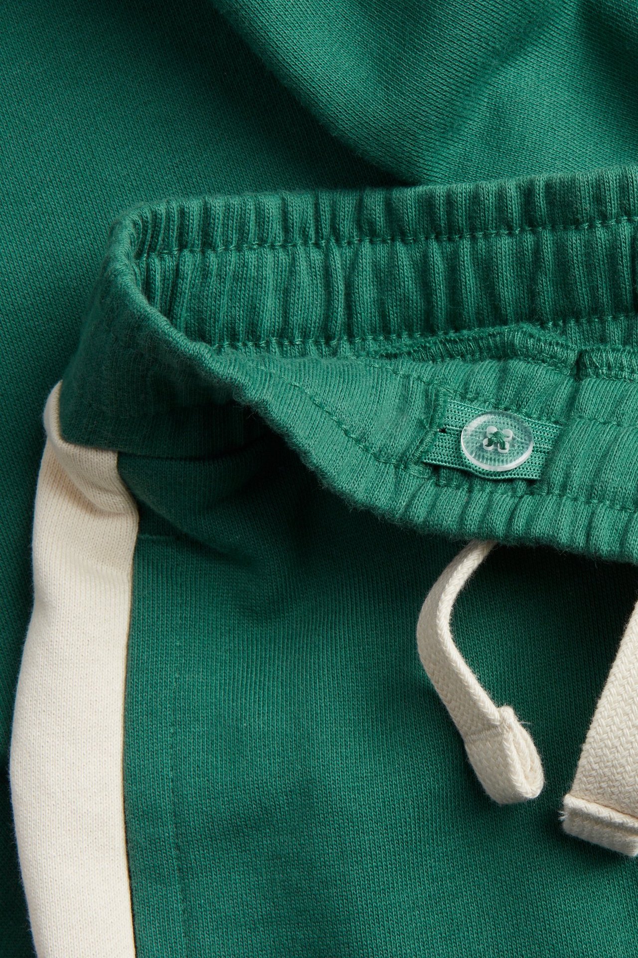 Zbliżenie zielonych bawełnianych spodni dresowych z elastycznym pasem, regulowanym guzikiem, sznurkiem i kremowym paskiem.