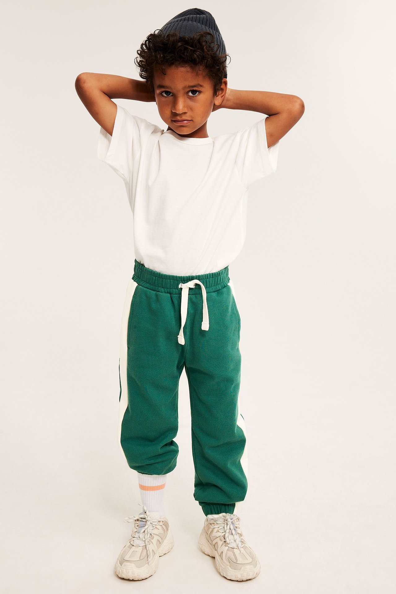 Barn i grønne bomullsjoggebukser med hvite sidestriper, en hvit t-skjorte og en mørkeblå lue.