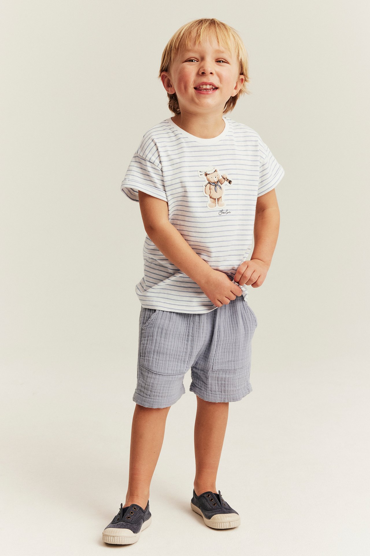Forfra, barn i offwhite stripet jerseyt-skjorte med bamse, lyseblå shorts og mørke sko.
