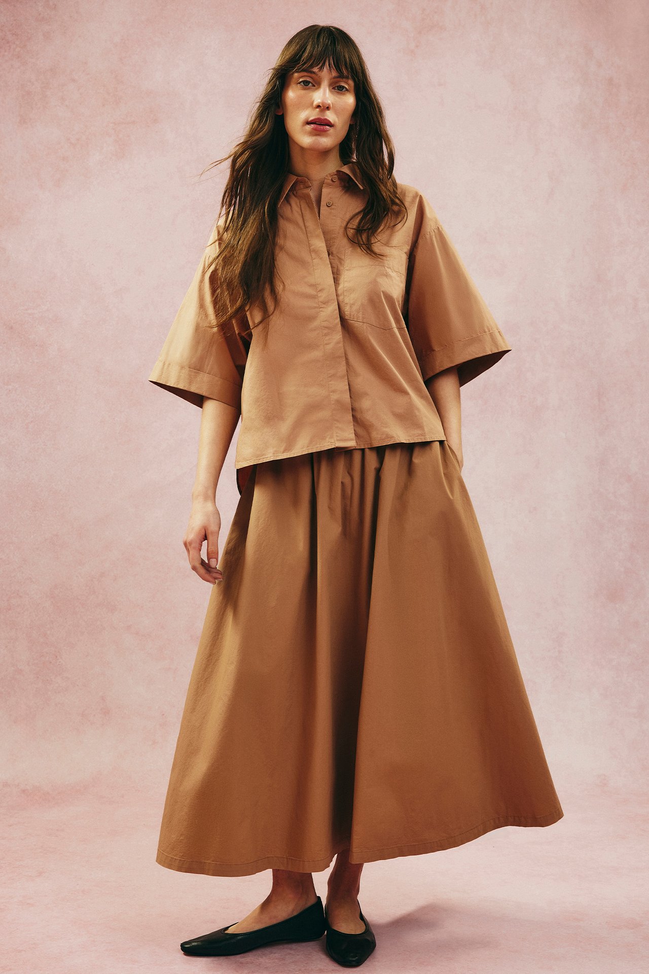 Framifrån, kvinna i beige utställd poplinkjol, matchande kortärmad skjorta och svarta ballerinaskor.