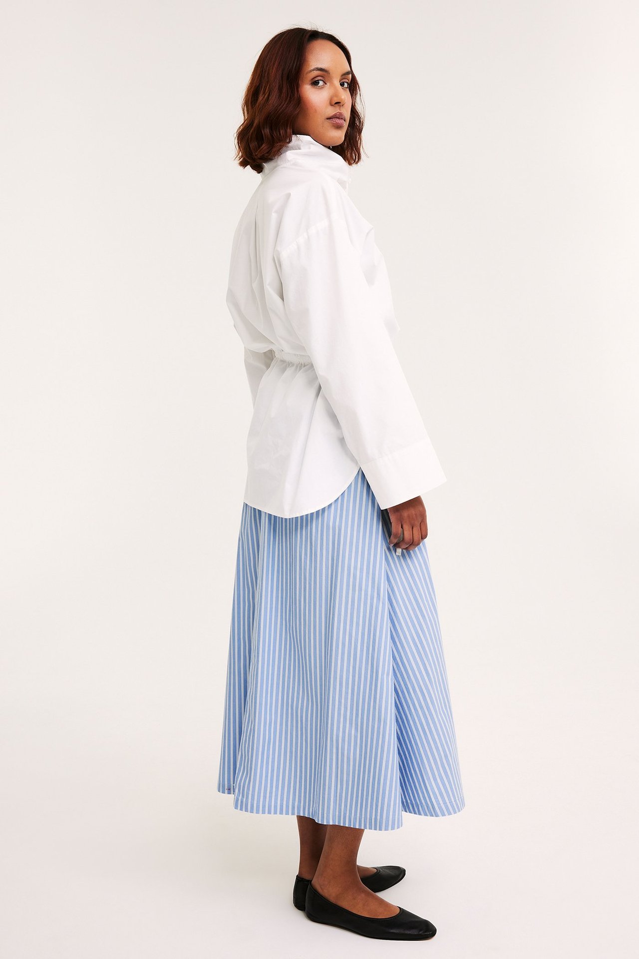 Sidestilt bilde av kvinne i lysblått stripet poplinskjørt, hvit oversized skjorte og sorte ballerinasko.