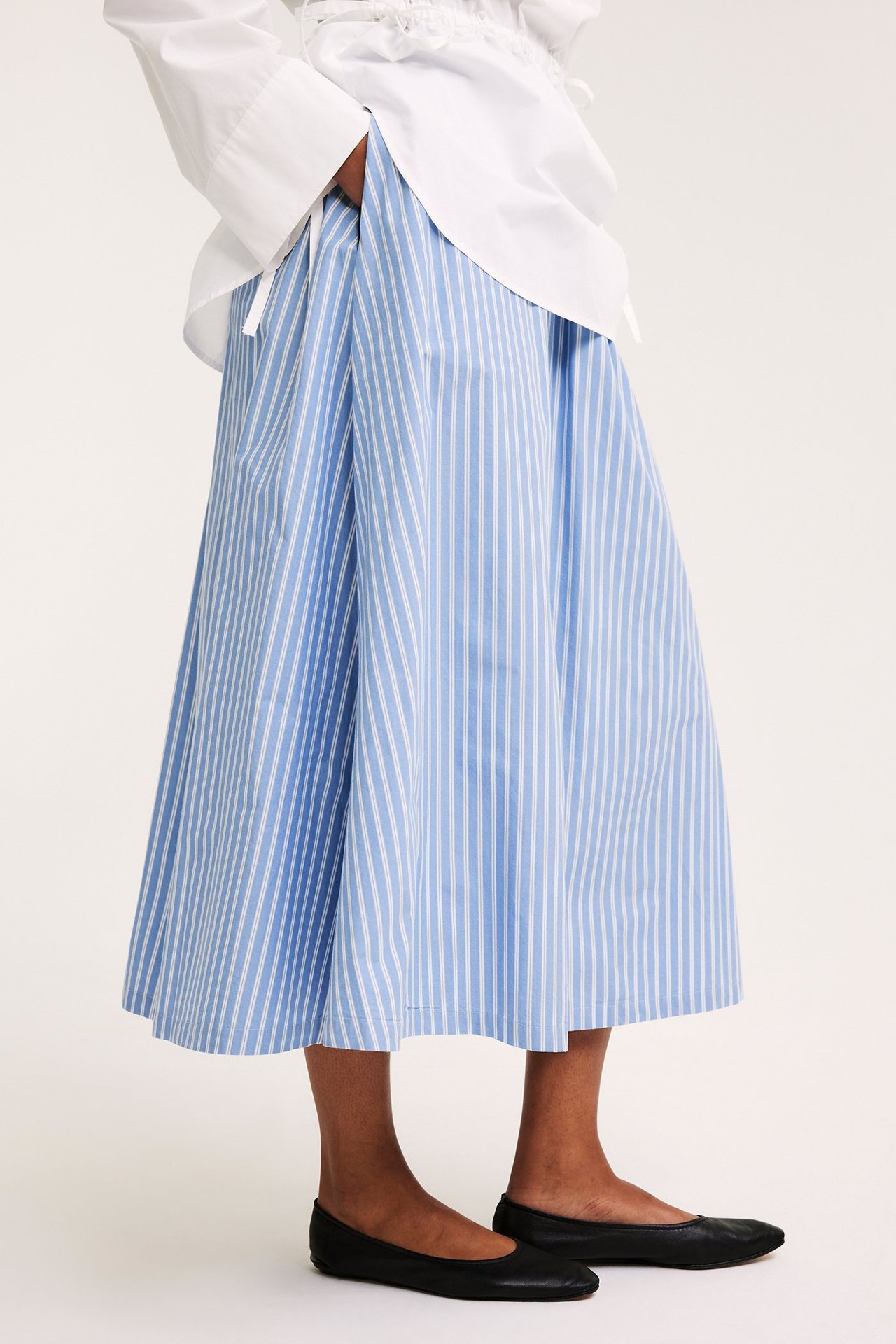 Sidebilde: kvinne i lyseblått stripete utsvunget poplinskjørt, hvit skjorte og svarte ballerinasko.