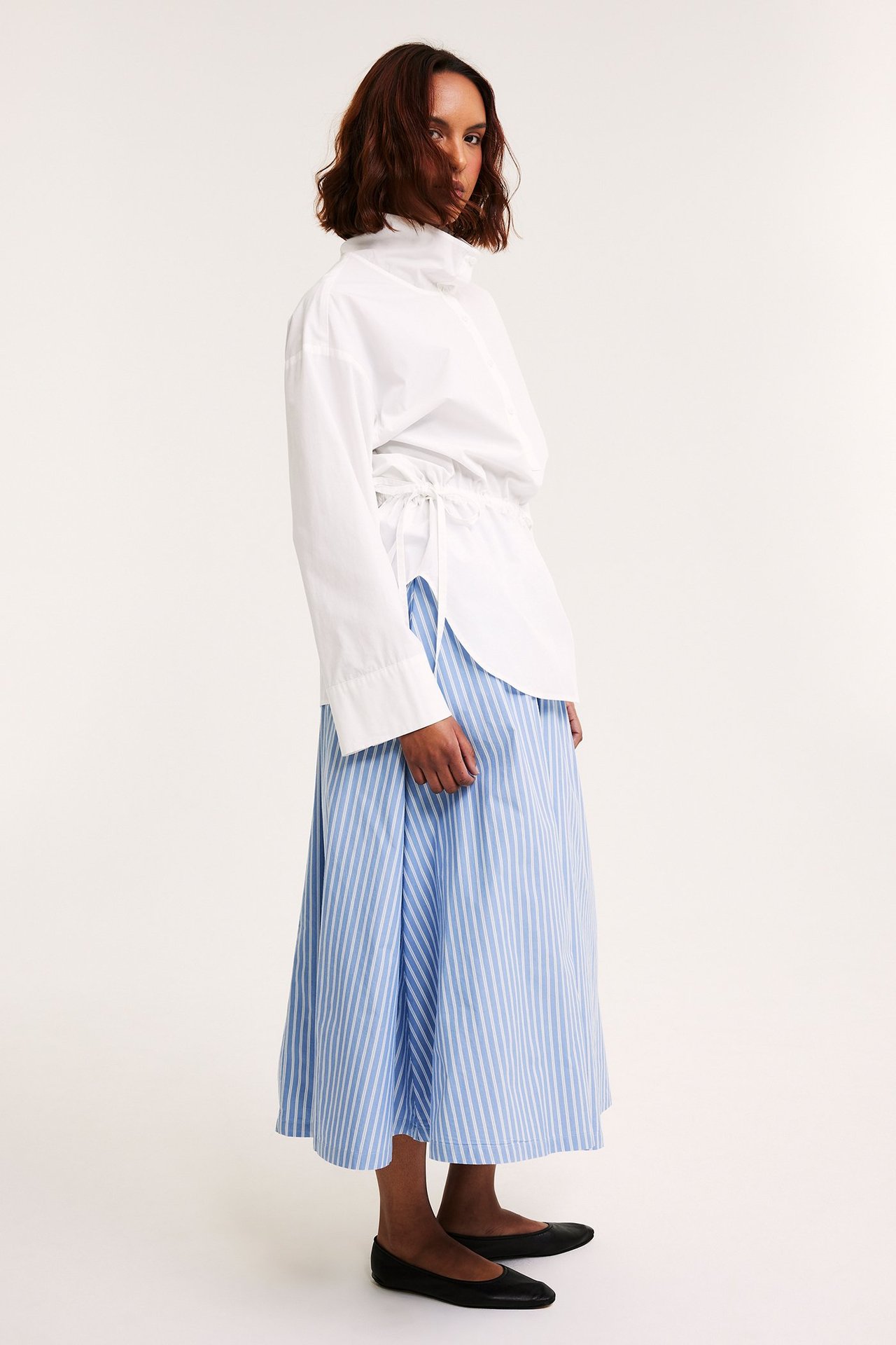 Sidebilde: Kvinne i lyseblåstripet utsvingt poplinskjørt, hvit bluse med snøring og svarte flate sko.