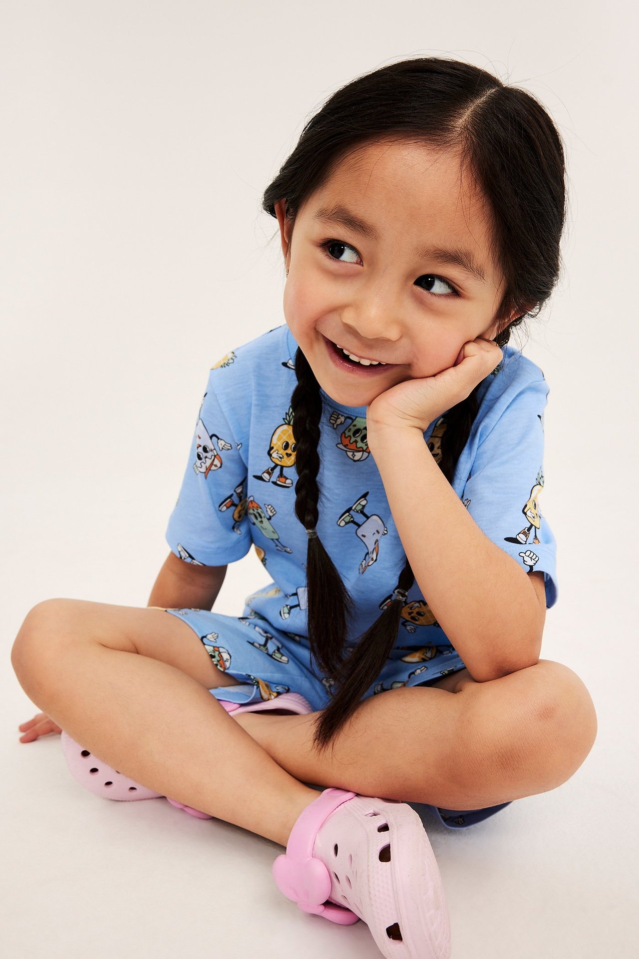 Sittande barn i halvprofil klädd i ljusblå mönstrad kortärmad pyjamas i bomull och rosa träskor.