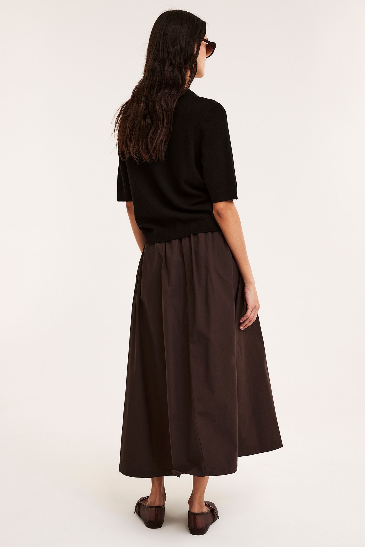 Bakifrån: kvinna i mörkbrun utställd bomullspoplinkjol, svart kortärmad topp och mörkbruna platta skor.