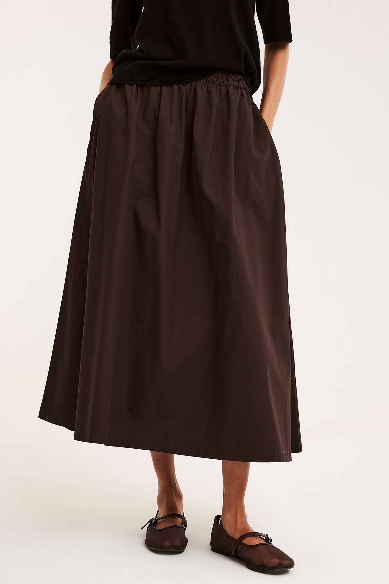 Frontbild av kvinna i mörkbrun utställd kjol i bomullspoplin, svart topp och nätskor.