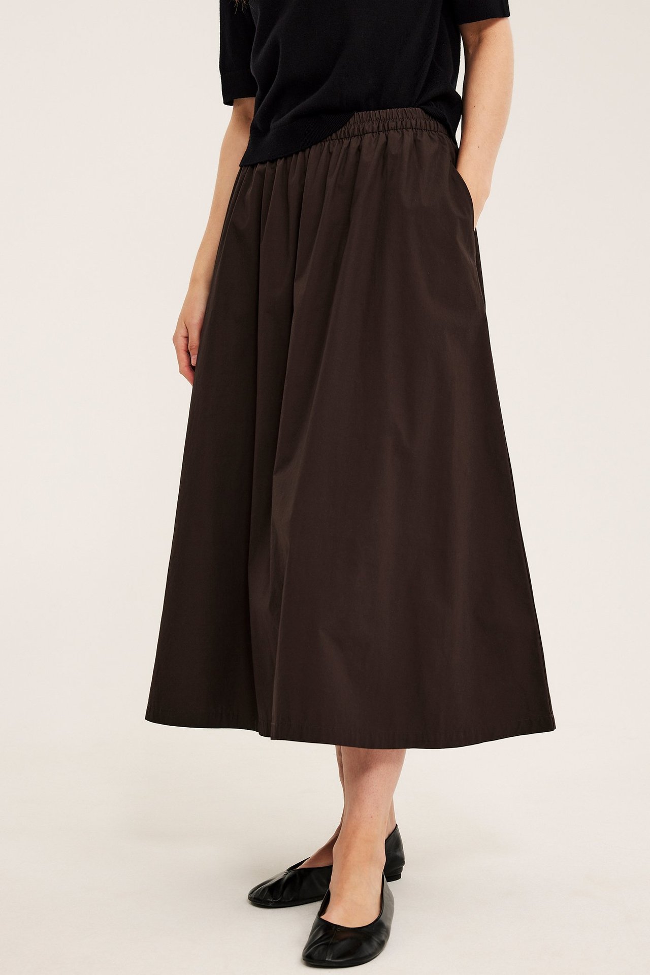 Näkymä edestä: nainen yllään tummanruskea levenevä puuvillapopliinihame, musta toppi ja mustat ballerinat.