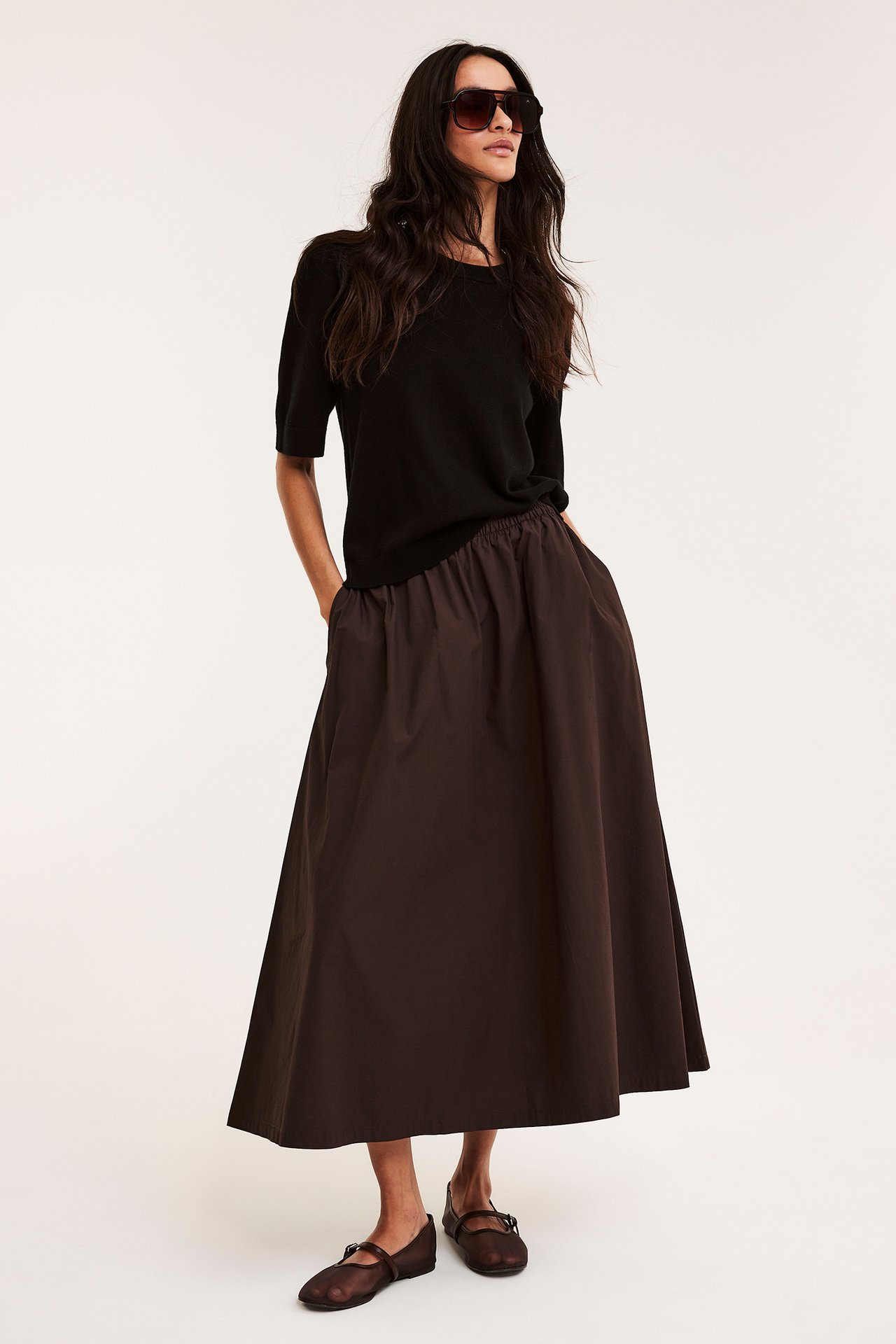 Front view: Kvinna i mörkbrun utställd poplinkjol, svart topp, bruna lågskor och solglasögon.