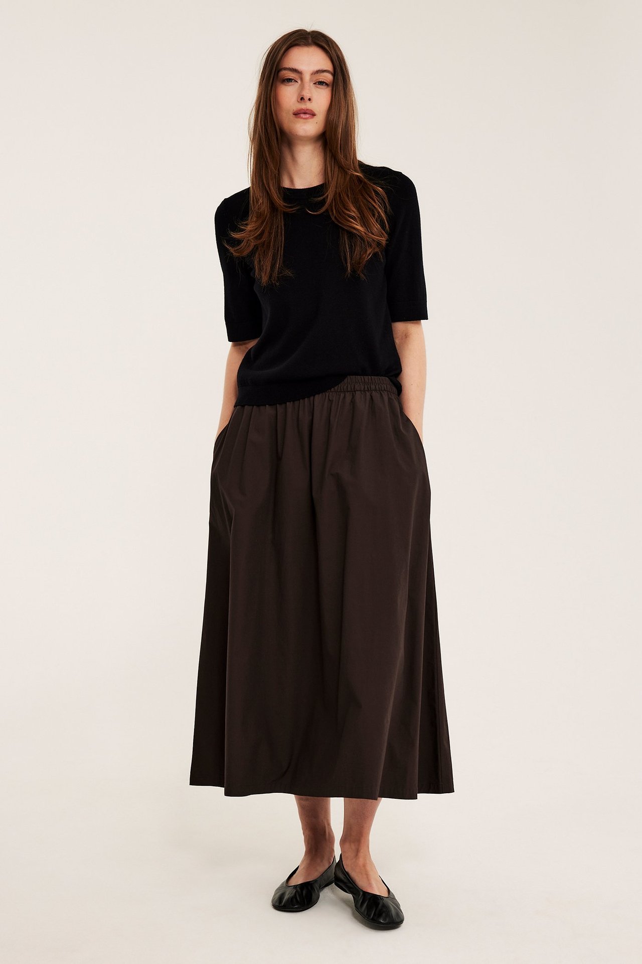 Nainen edestä tummanruskeassa levenevässä puuvillapopliinihameessa, mustassa lyhythihaisessa topissa ja mustissa ballerinoissa.