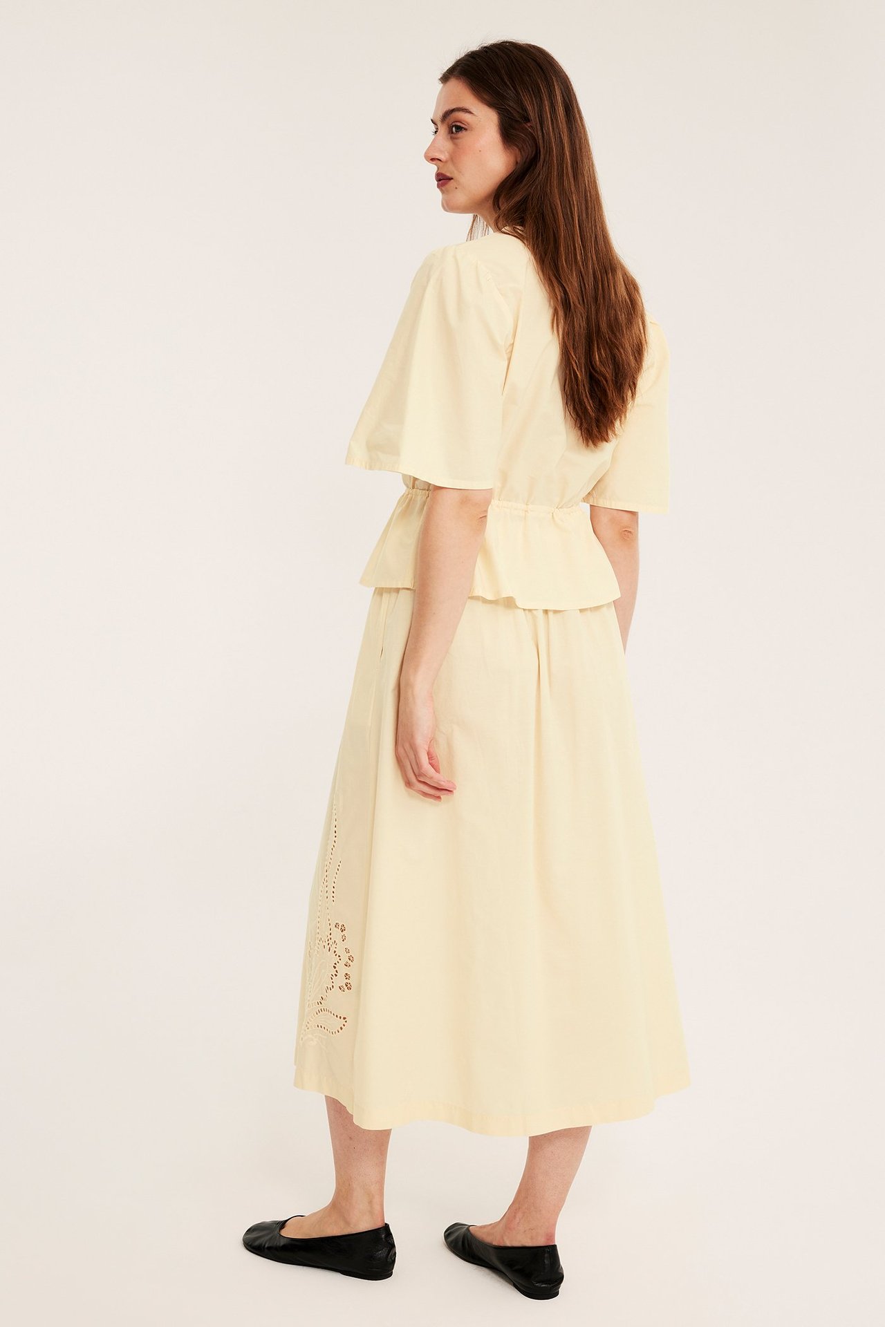 Taka-sivukuva: Nainen vaaleankeltaisessa brodeeratussa popliinihameessa, yhteensopiva toppi ja mustat kengät.