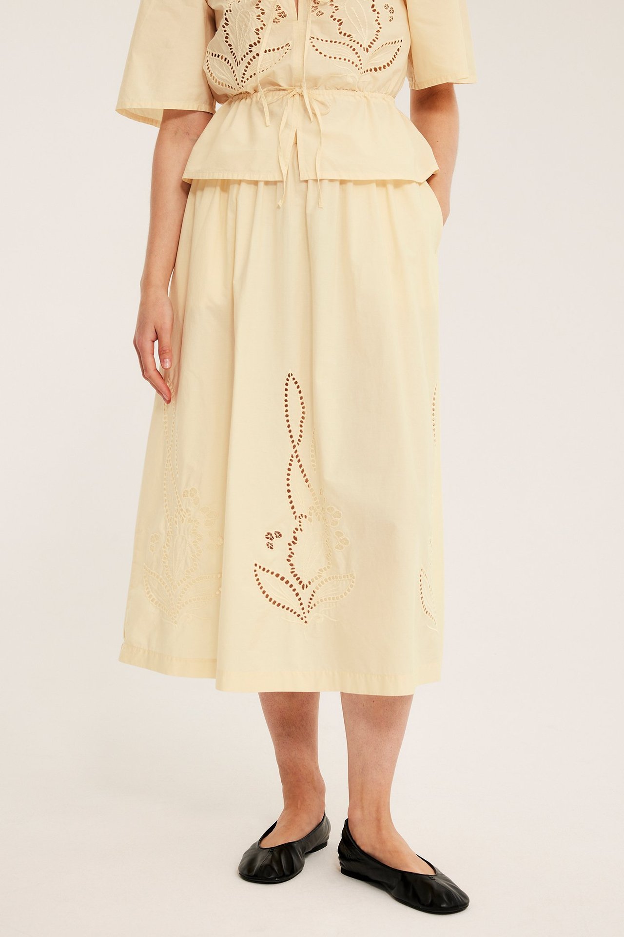 Edestä: Nainen vaaleankeltaisessa brodeeratussa leveässä puuvillahameessa, yhteensopivassa topissa, mustissa ballerinoissa.