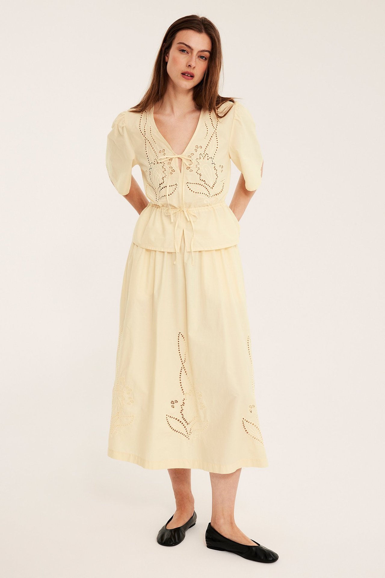 Edestäpäin: nainen yllään leveä vaaleankeltainen brodeerattu puuvillapopliinihame, yhteensopiva peplum-pusero ja mustat ballerinakengät.