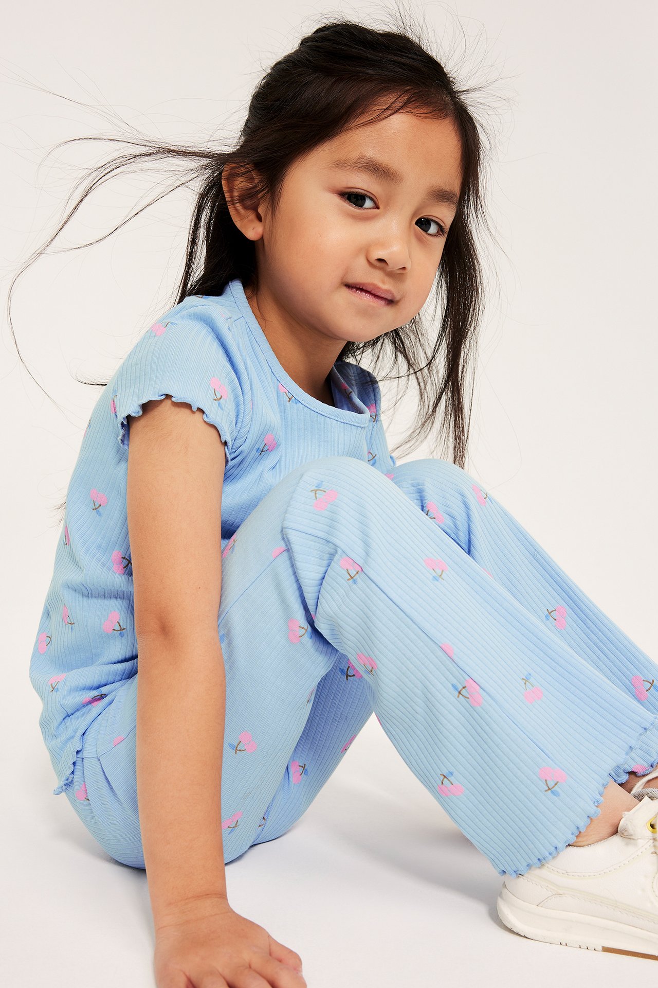 Forfra en kid i lyseblå ribbestrikkede flared leggings med rosa kirsebærtrykk og en matchende kortermet topp.