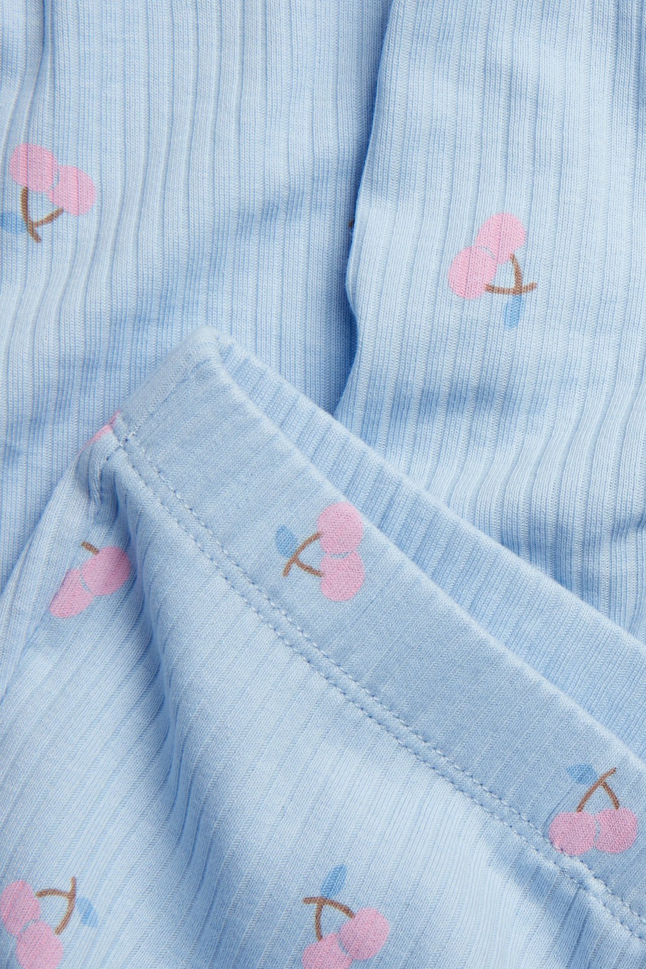 Lähikuva vaaleansinisistä ribatuista flare-leggingseistä vaaleanpunaisella kirsikkakuviolla ja kuminauhavyötäröllä.