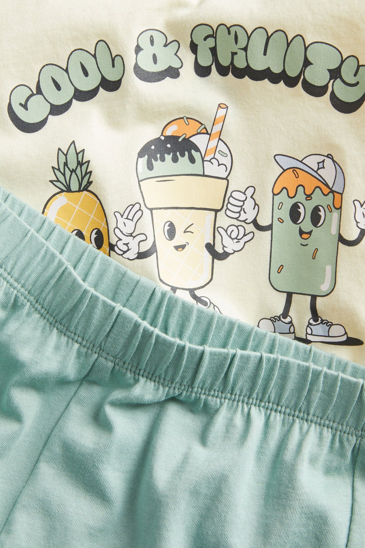 Zbliżenie na dziecięcą jasnożółtą bawełnianą piżamę z krótkim rękawem, nadrukiem owoców i jasnozielone spodenki.