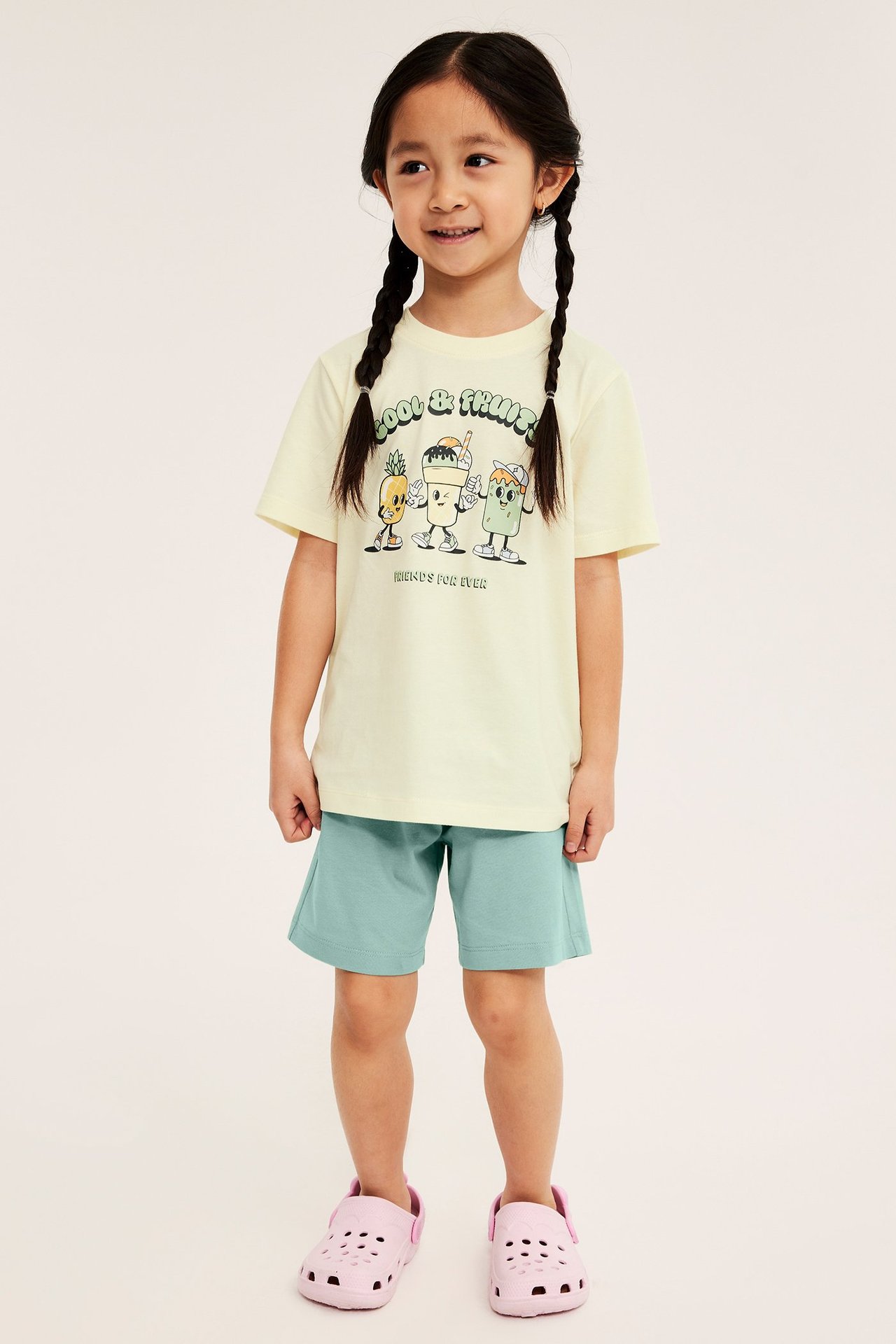 Frontvy: Kid i ljusgul kortärmad pyjamas med frukttryck på tröjan och ljusgröna shorts.