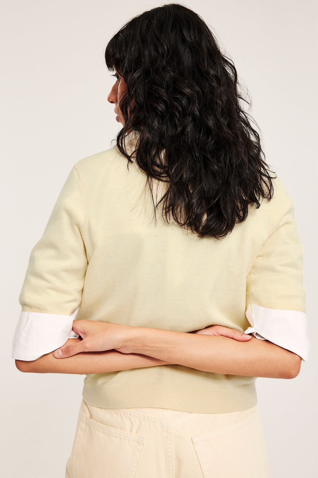 Kobieta od tyłu: jasnożółty krótki sweter z mieszanki wełny, na białej koszuli i jasnobeżowych spodniach.