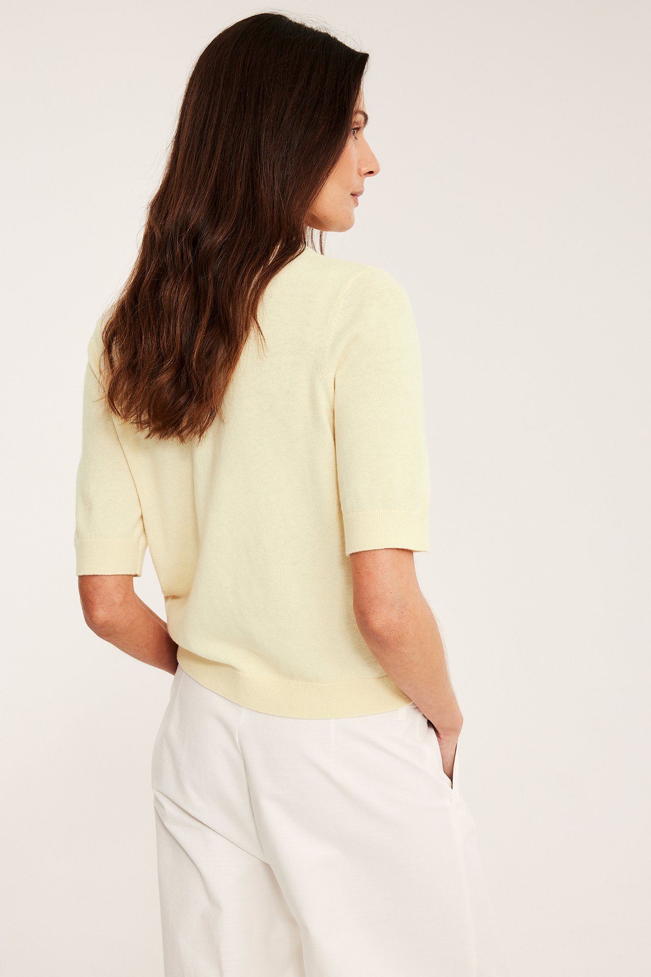 Widok z boku i tyłu: Kobieta w jasnożółtym swetrze z krótkim rękawem z wełny i białych spodniach.