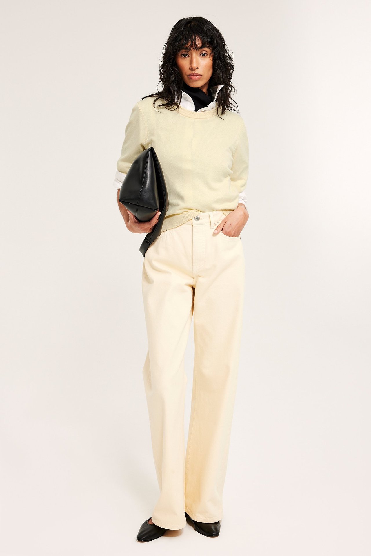 Widok z przodu. Kobieta nosi jasnożółty wełniany sweter na białej koszuli, jasnożółte spodnie, trzyma czarną kopertówkę.