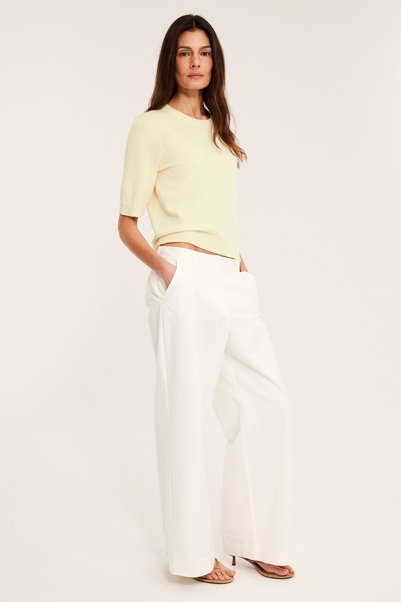 Forfra: kvinne i lys gul kortermet finstrikket genser, hvite vide bukser og beige sandaler.