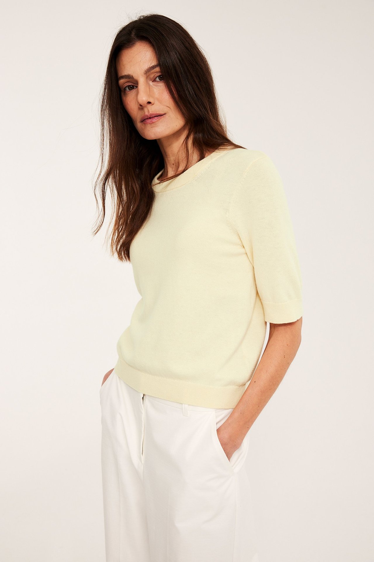 Widok z przodu: Kobieta w jasnożółtym, krótko rękawowym, drobno dzianym swetrze z wełny i białych spodniach.