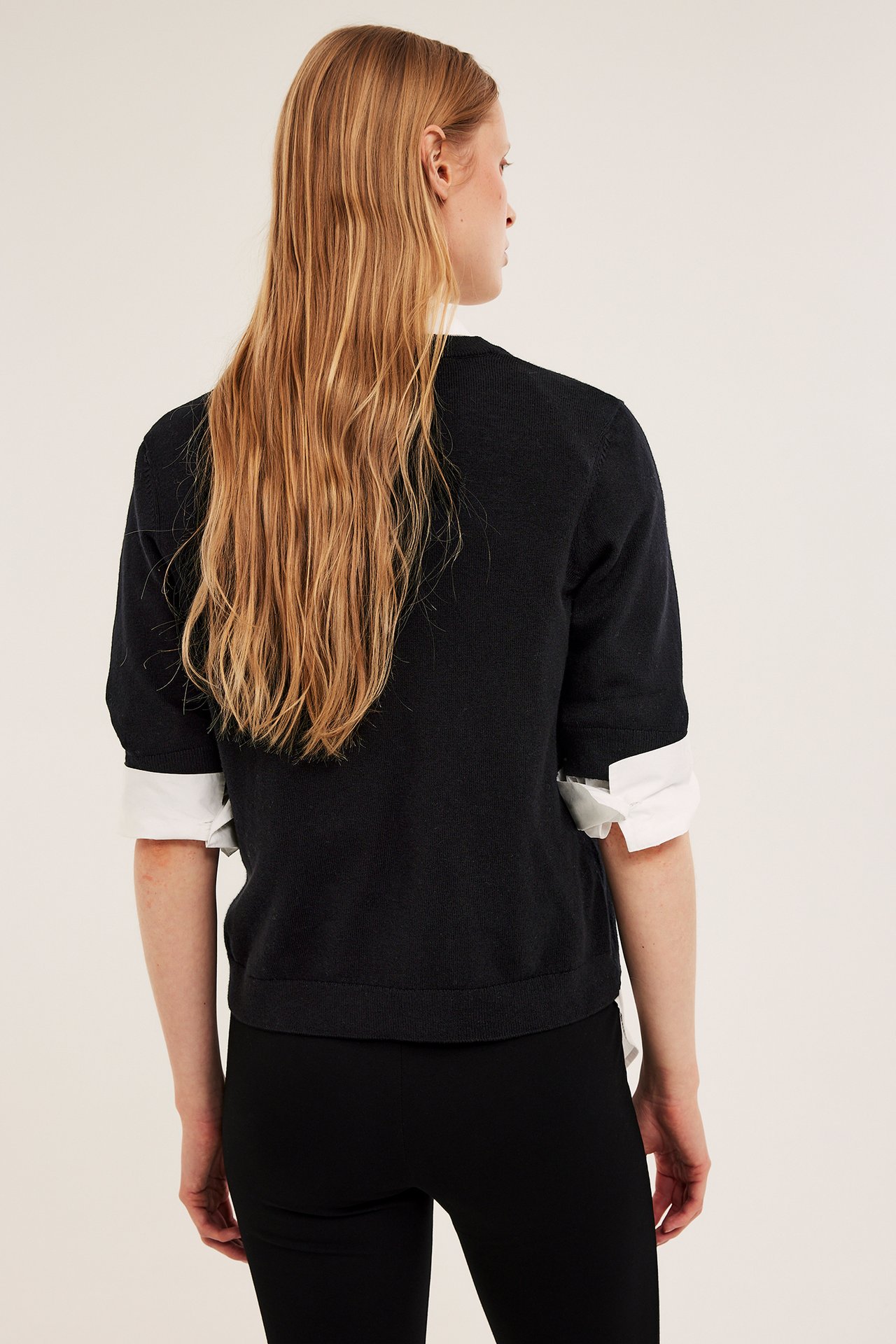 Widok z tyłu kobiety w czarnym, wełnianym swetrze z krótkim rękawem, na białej koszuli i czarnych spodniach.
