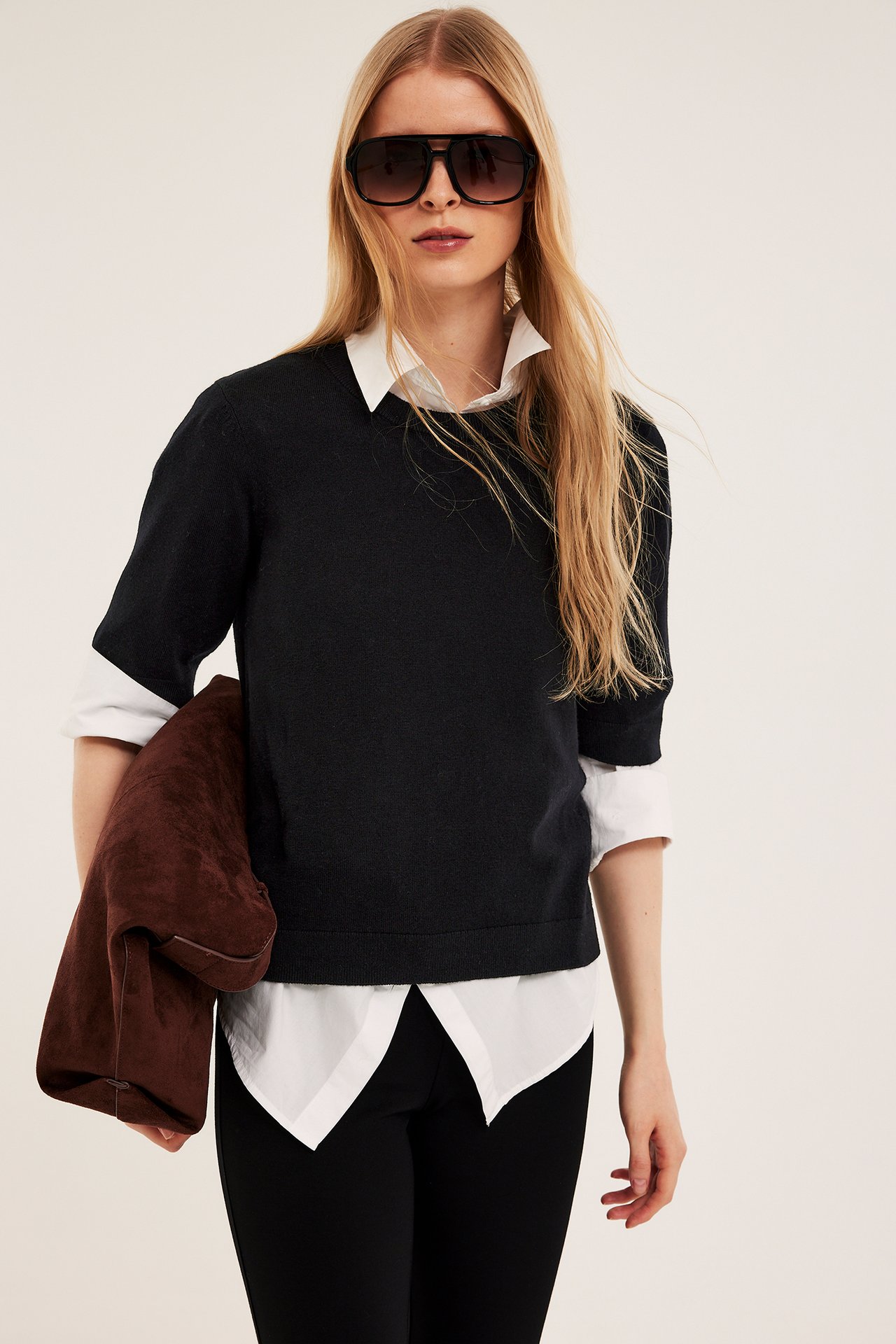 Kobieta w czarnym krótkim swetrze z mieszanki wełny na białej koszuli, czarnych spodniach, okularach, trzyma brązową kurtkę.