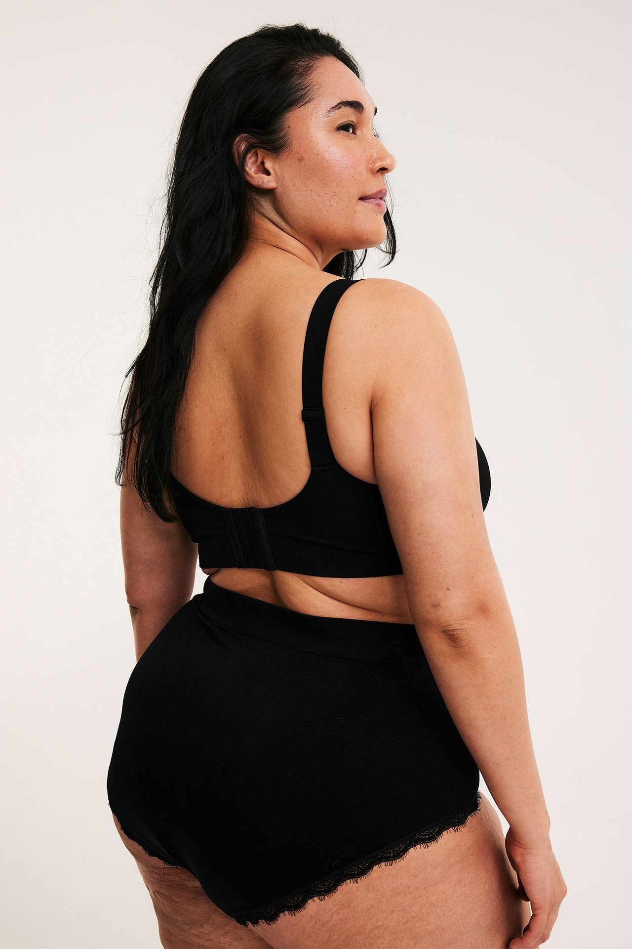 Kvinna bakifrån, i en svart sömlös bygellös bh med hyskknäppning och svarta spetskantade trosor.