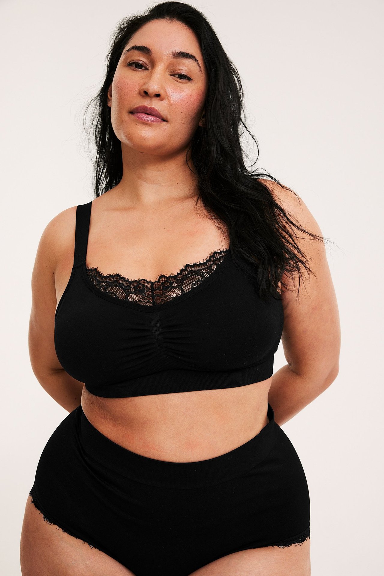 Frontvy: Kvinna bär en svart sömlös bygellös bh med spetskant, rynk framtill, och matchande trosa.